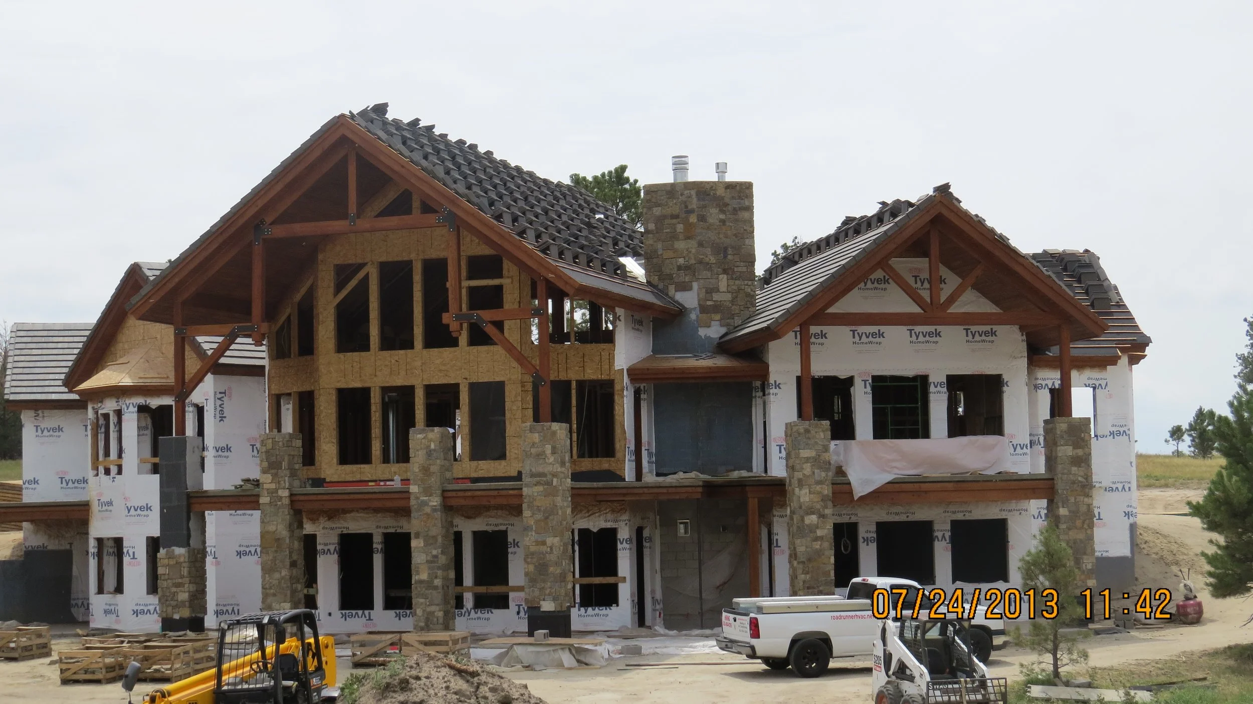 Under-construction house with wooden framework, stone chimney, and partially installed roofing, surrounded by construction equipment and vehicles, with a cloudy sky.