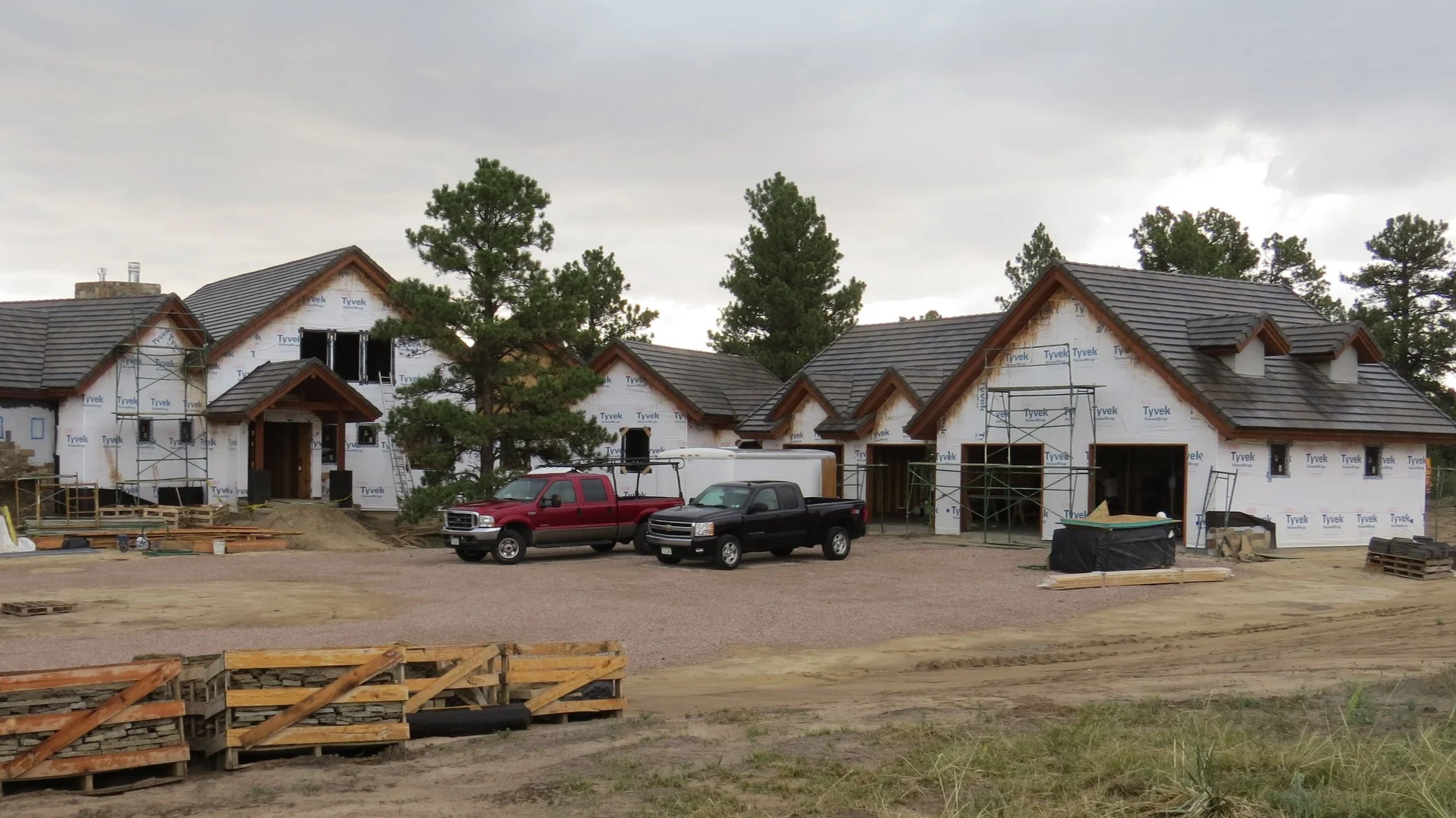 Construction site of a residential neighborhood with multiple houses in progress, scaffolding and vehicles parked in front, surrounded by trees and cloudy sky.
