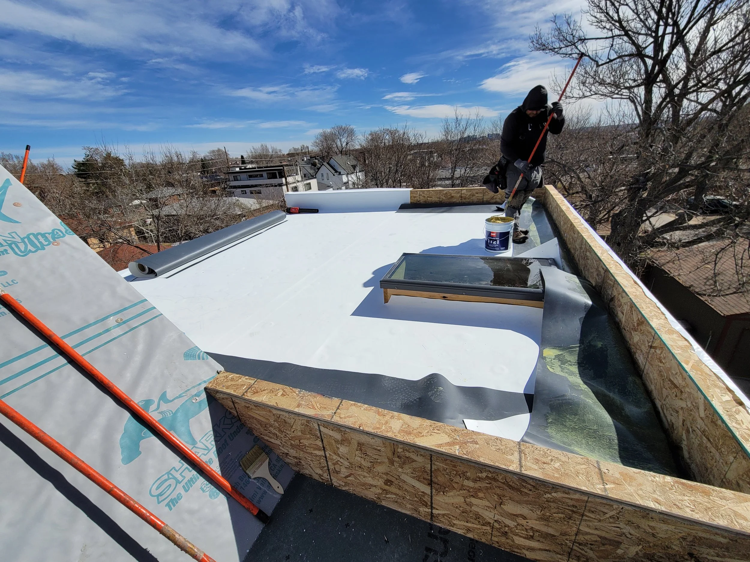 A worker on a rooftop installing roofing materials. The sky is blue with some clouds, and there are trees and buildings in the background.