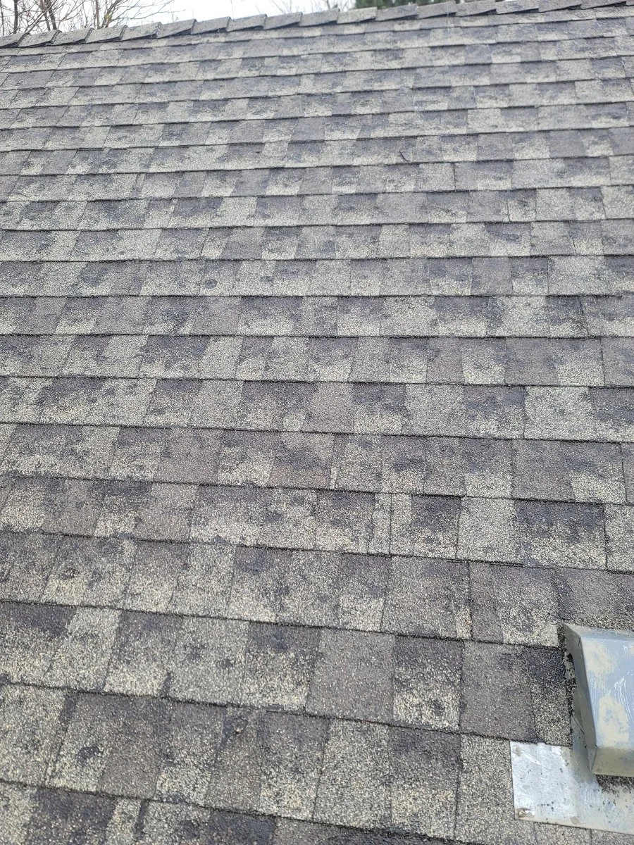 A close-up view of a shingled rooftop with asphalt shingles in gray and black tones, showing damage caused by hail impacts.