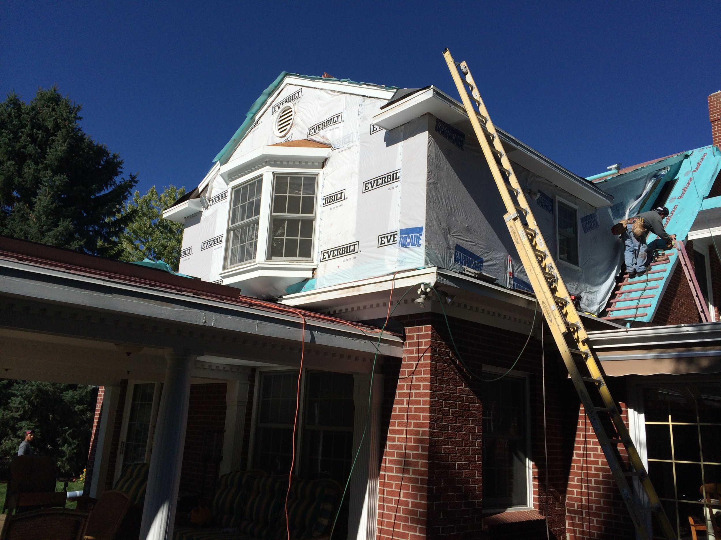 Construction workers installing or repairing the roofing of a brick house, with a ladder and building materials visible under a clear blue sky.