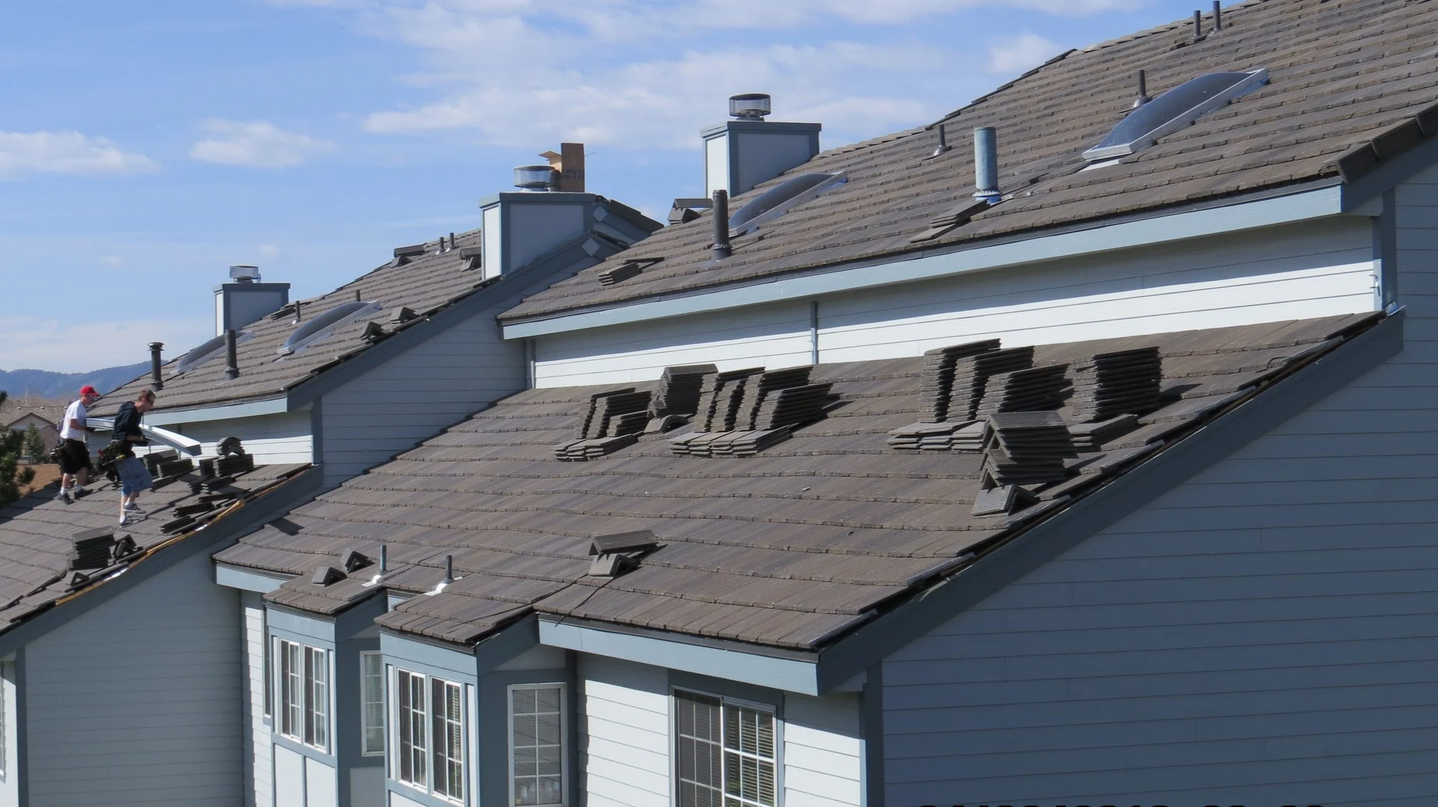 Two workers on a house roof, installing new shingles with stacks of shingles nearby, under a blue sky.