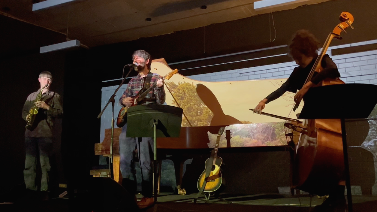 Three musicians performing on stage: a saxophonist, a guitarist singing into a microphone, and a cellist adjusting her instrument. There is a guitar on the floor and a scenic projection behind them.