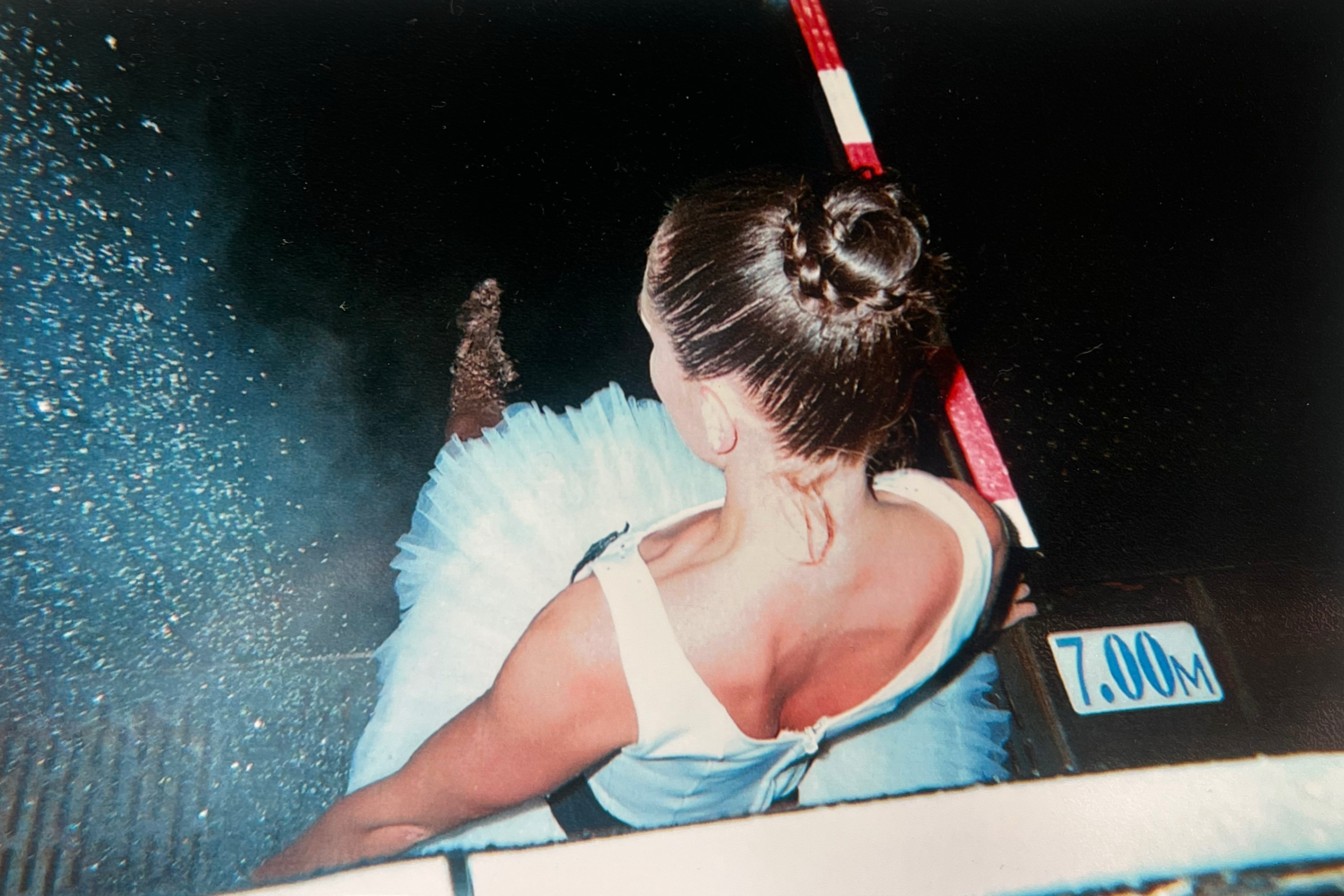 A ballet dancer in a white tutu performing on stage with a dark background. The image is taken from behind, showing the dancer's hair in a bun and her arm extended.