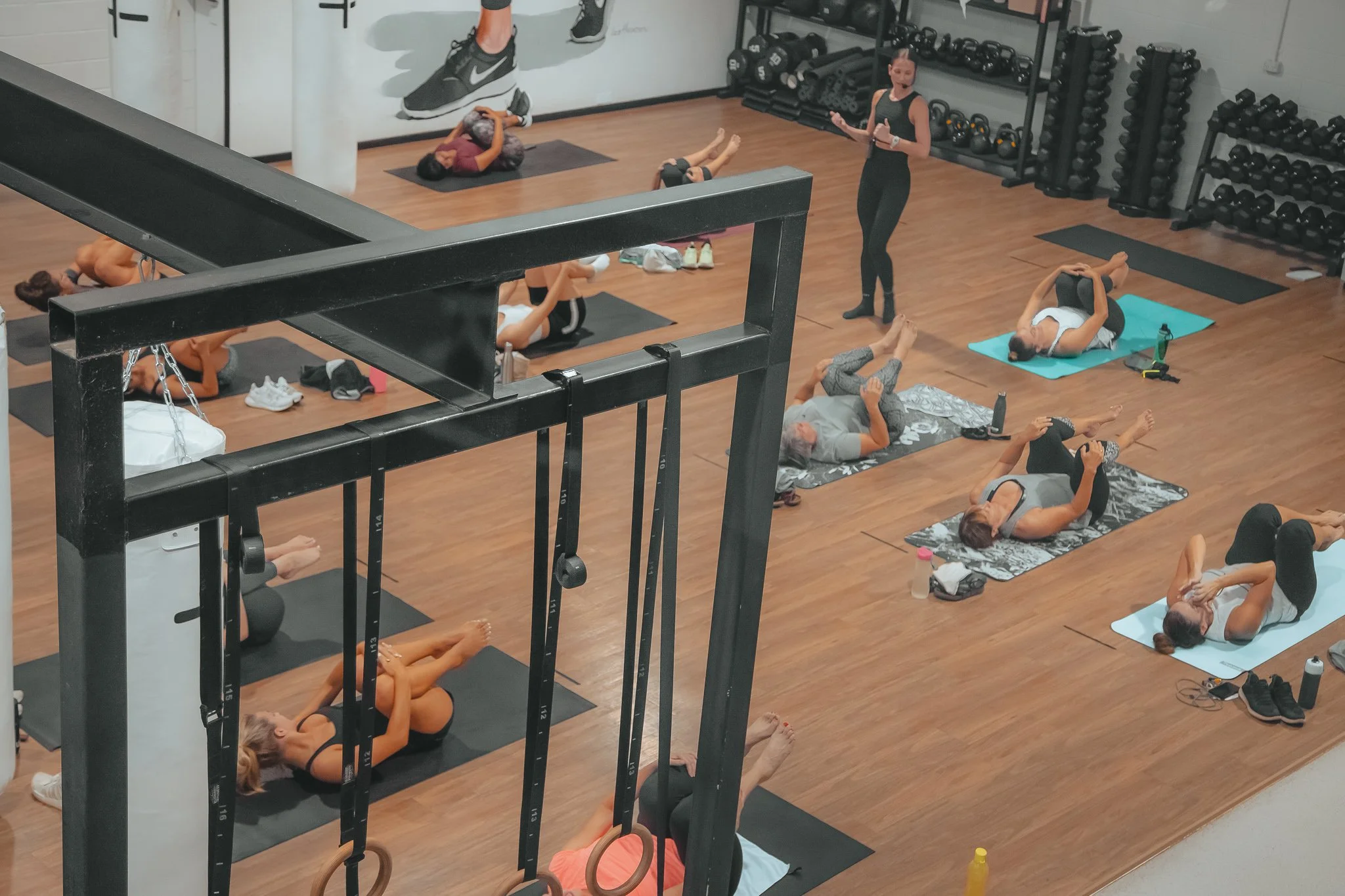 Female instructor - Katie Baring-Gould coaching a Pilates Sculpt class on mats in a gym. The instructor is standing and giving instructions, surrounded by exercise equipment.