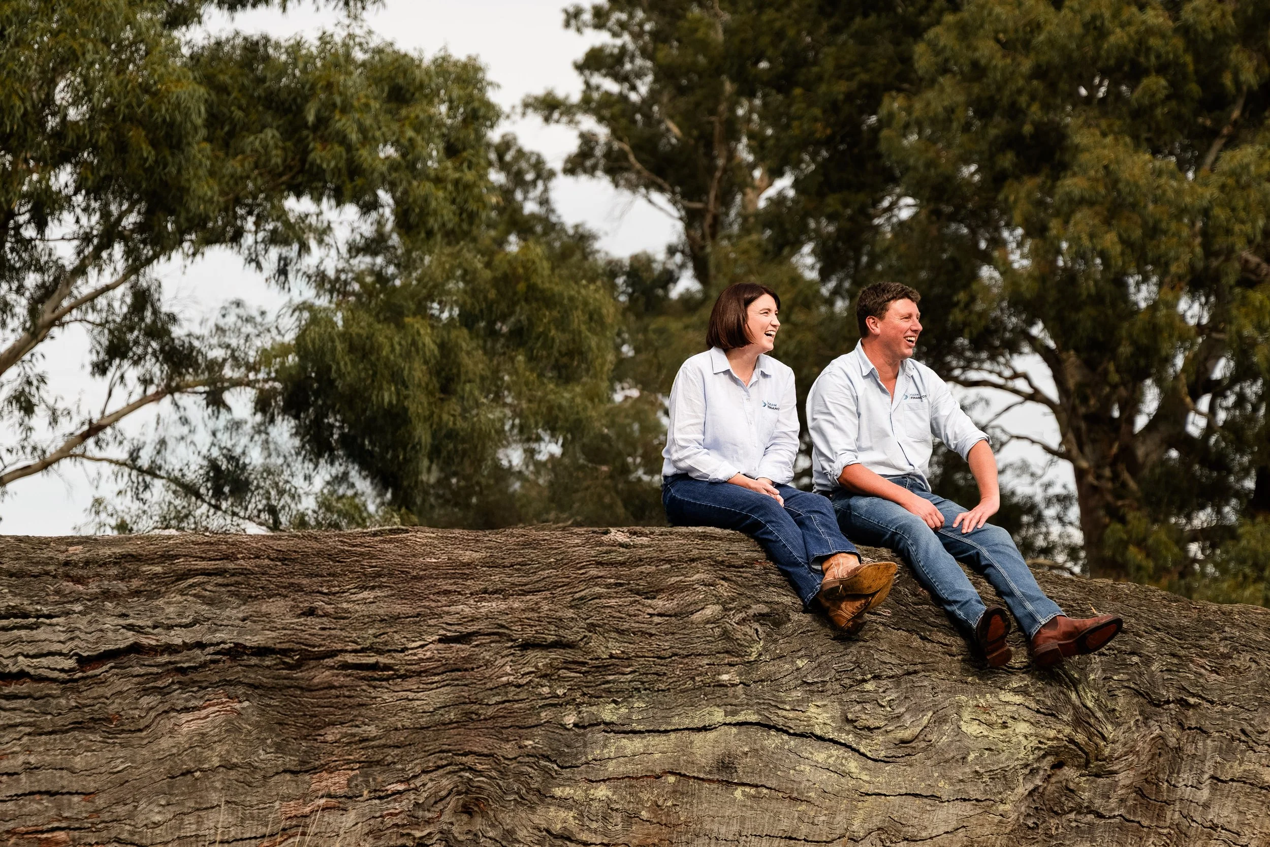 Members of Farm Finance Co Lachie and Alana, sitting on a large fallen tree trunk outdoors, smiling and laughing, with trees and a cloudy sky in the background.