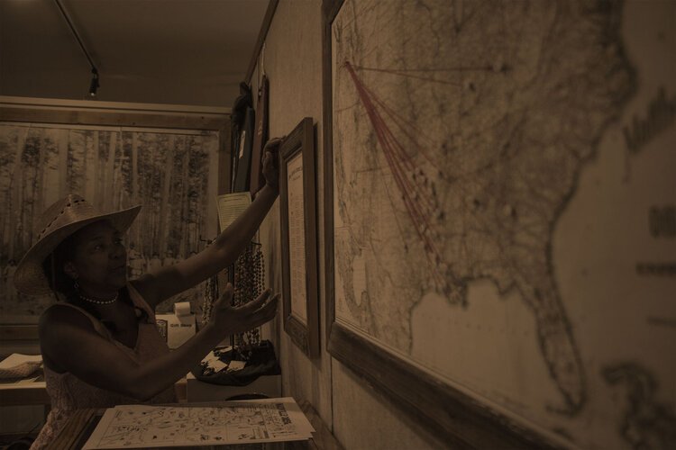A woman wearing a wide-brimmed straw hat and pearl necklace, reaching out to a framed picture on a wall with maps in the background.