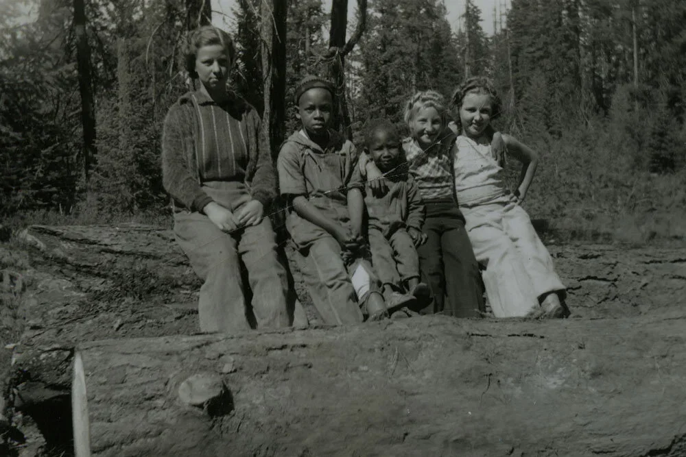 Black and white photo of five children sitting on a log in a forest setting.