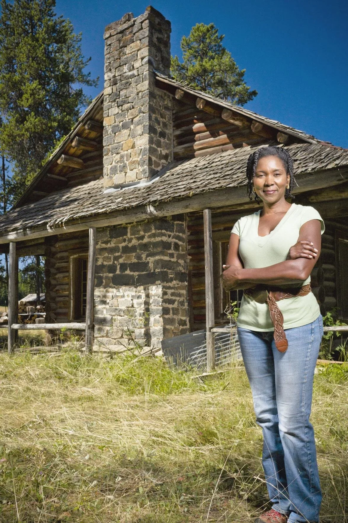 Woman standing outdoors in front of an old log cabin with a stone chimney, crossed arms, smiling, with trees and blue sky in the background.