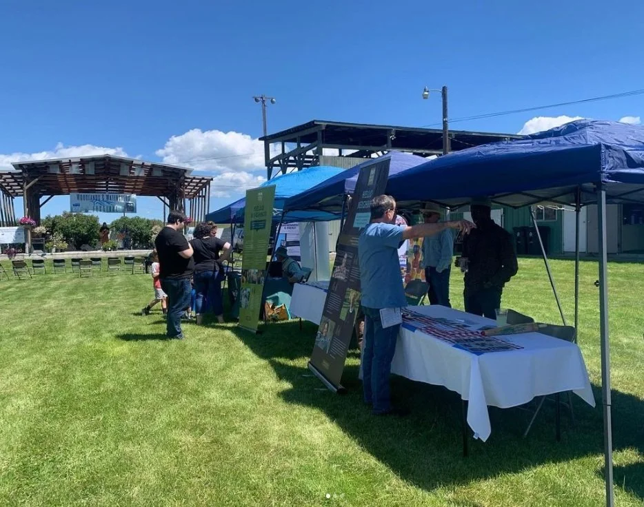 People attending an outdoor event under blue tents on a grassy area, with informational banners and tables set up, and a wooden stage in the background under a blue sky with some clouds.