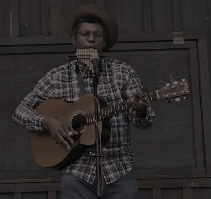 A man wearing a plaid shirt and hat playing an acoustic guitar and singing into a microphone.