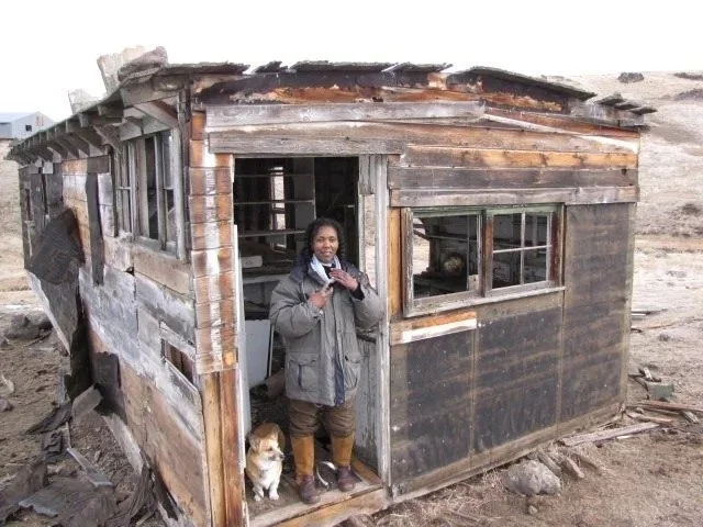 Person standing in front of a small, weathered wooden house with a dog nearby.