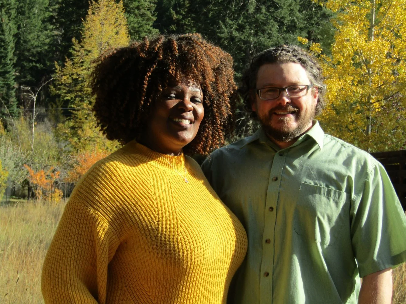 A woman wearing a yellow sweater and a man wearing glasses and a light green shirt stand outdoors with trees showing fall foliage in the background.