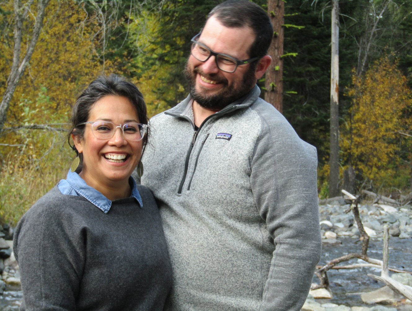 A smiling couple standing outdoors in a wooded area with autumn trees and a rocky stream in the background.