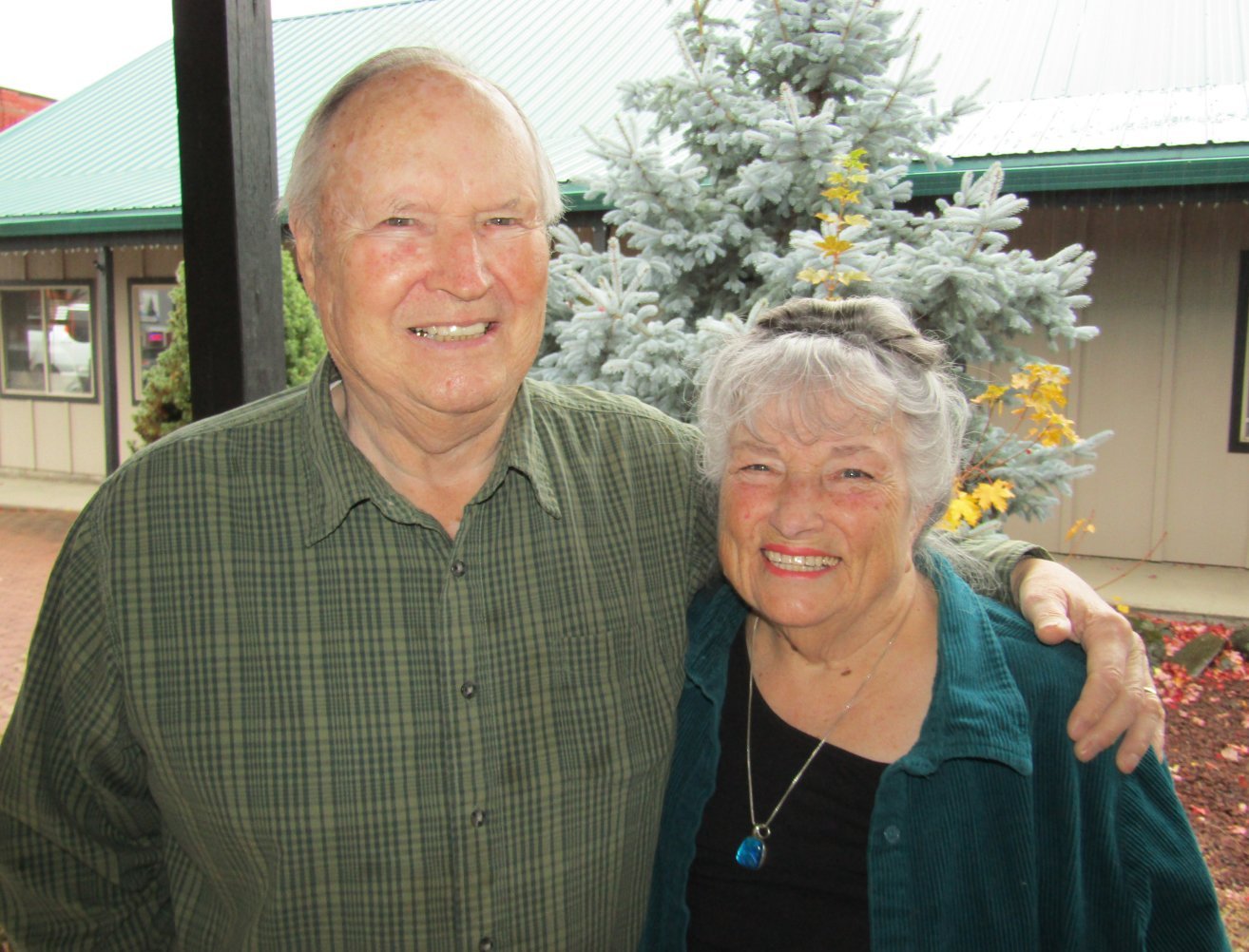 An elderly man and woman standing outdoors, smiling, with a blue spruce tree and a building in the background.