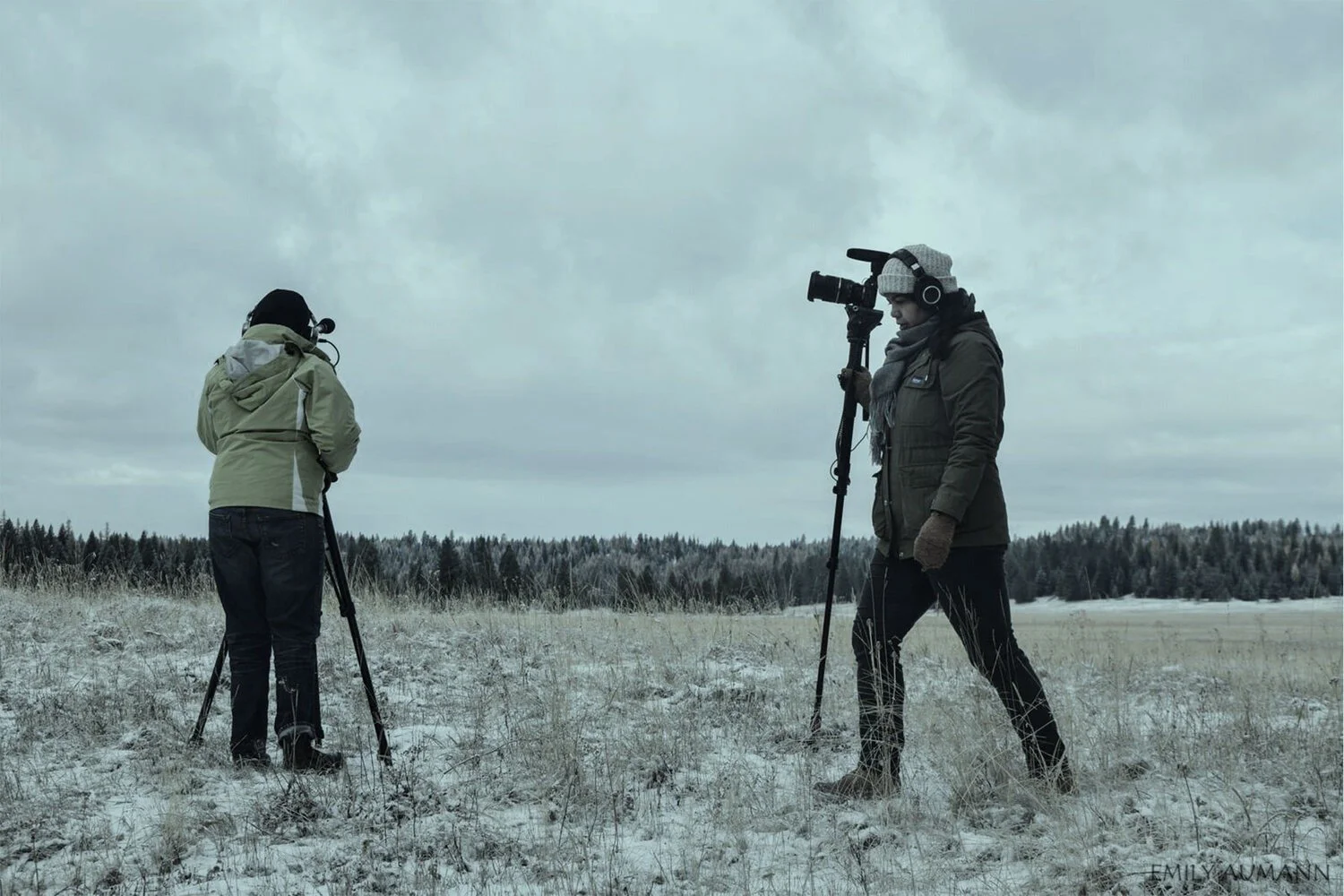 Two people filming outdoors in a snowy field with a forest in the background, overcast sky, one person is looking through a camera on a tripod and the other is holding a large camera with a shoulder strap.