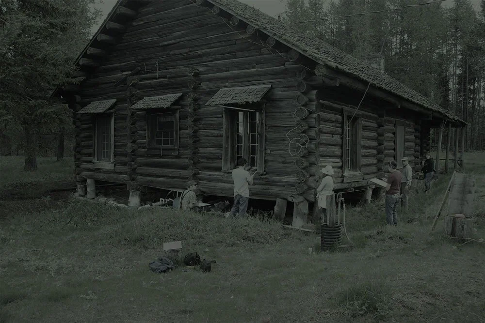 People working on an old wooden log cabin in a forested area.