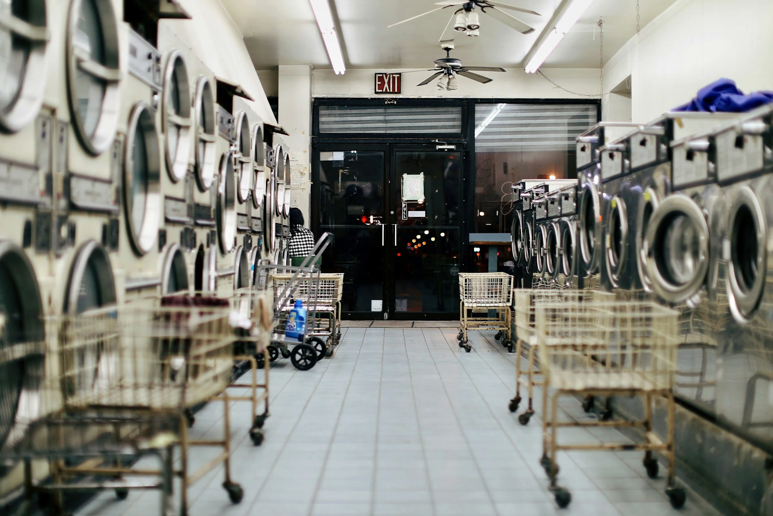 Laundry room with washing machines on both sides, shopping carts in the aisle, ceiling fans, and an exit door at the back.