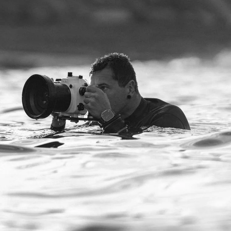 A man in water, looking through a camera or scope, with a black wristwatch, on a body of water during daylight.