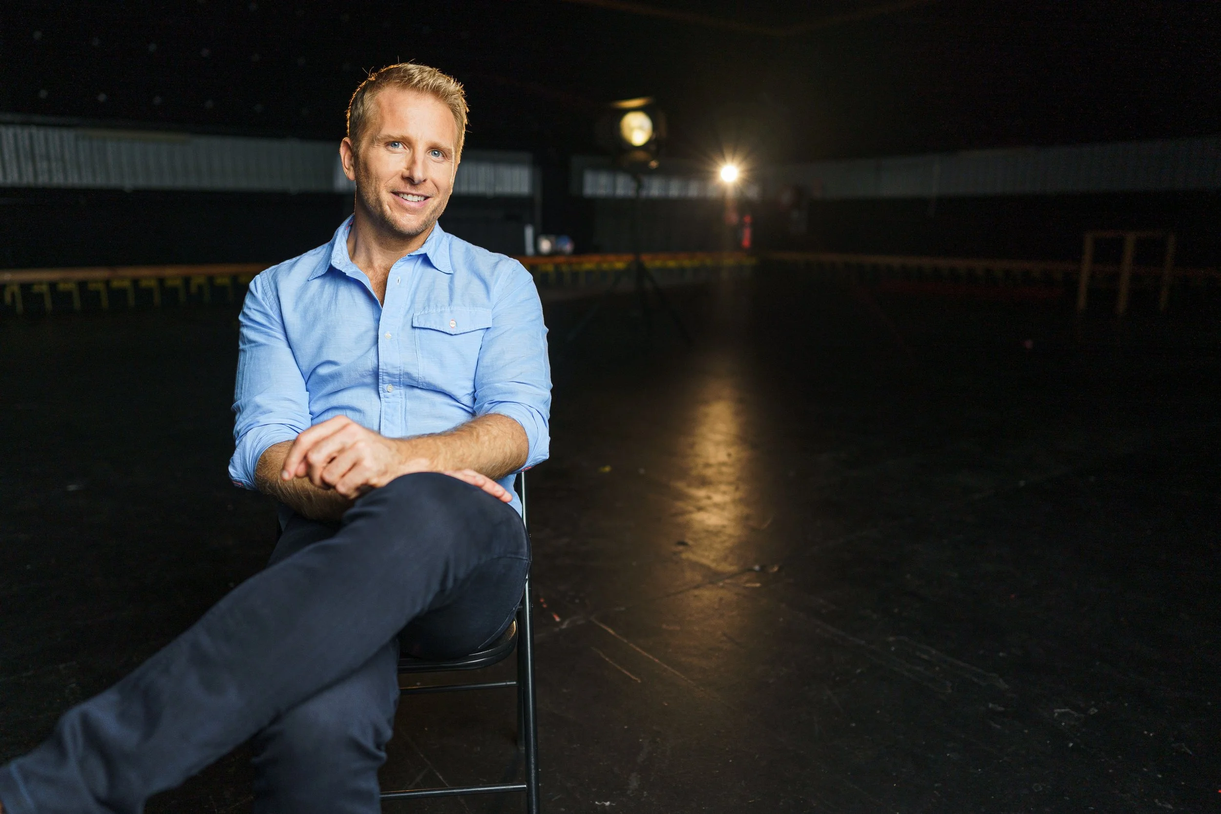 A man with light hair and fair skin, dressed in a light blue shirt, sitting in a chair in a dark space with a lit spotlight in the background.