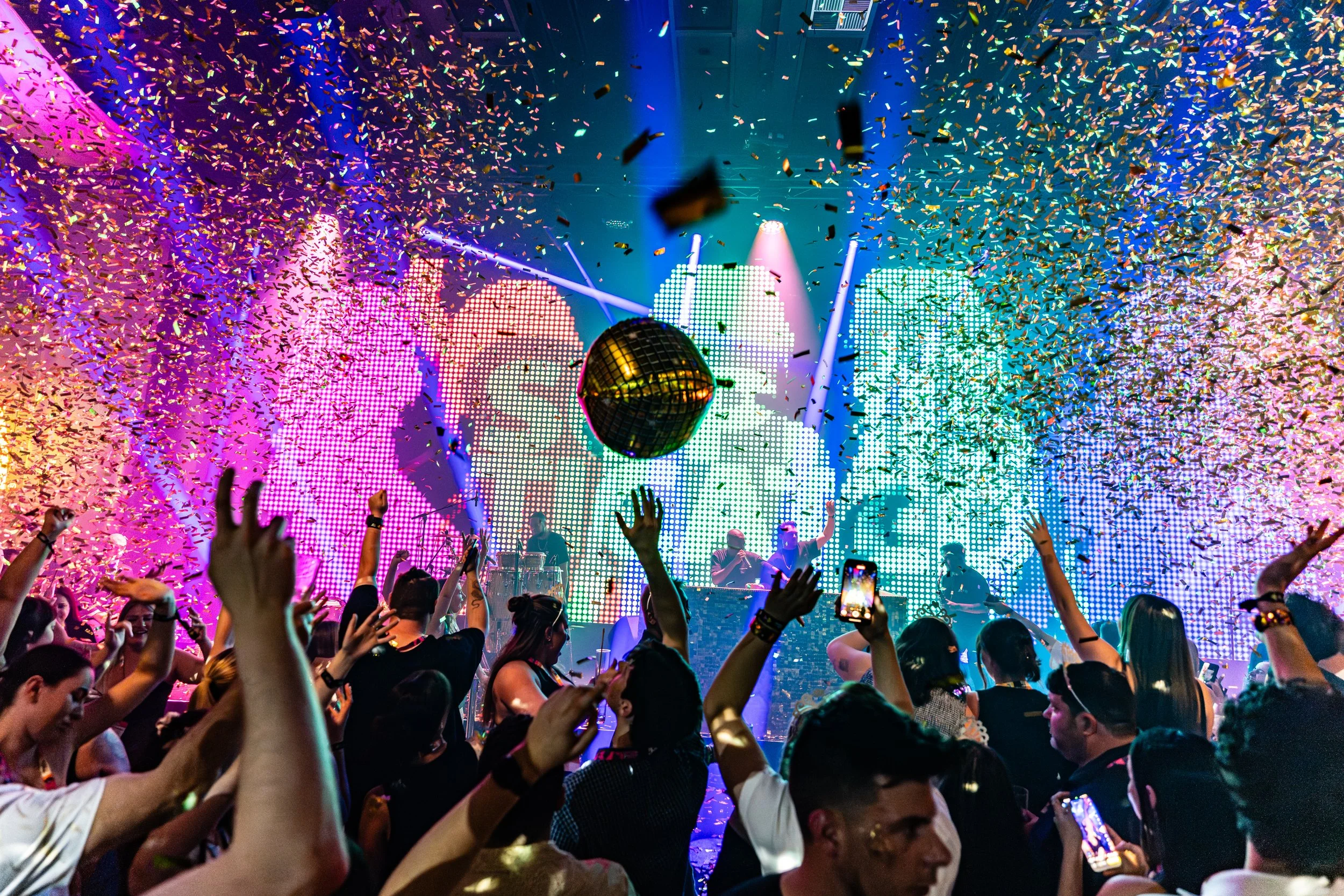 A lively concert or nightclub scene with a crowd of people dancing and enjoying the party. The background features colorful LED lights, a DJ or performers on stage, and a disco ball hanging from the ceiling. Confetti is falling from above, adding to 