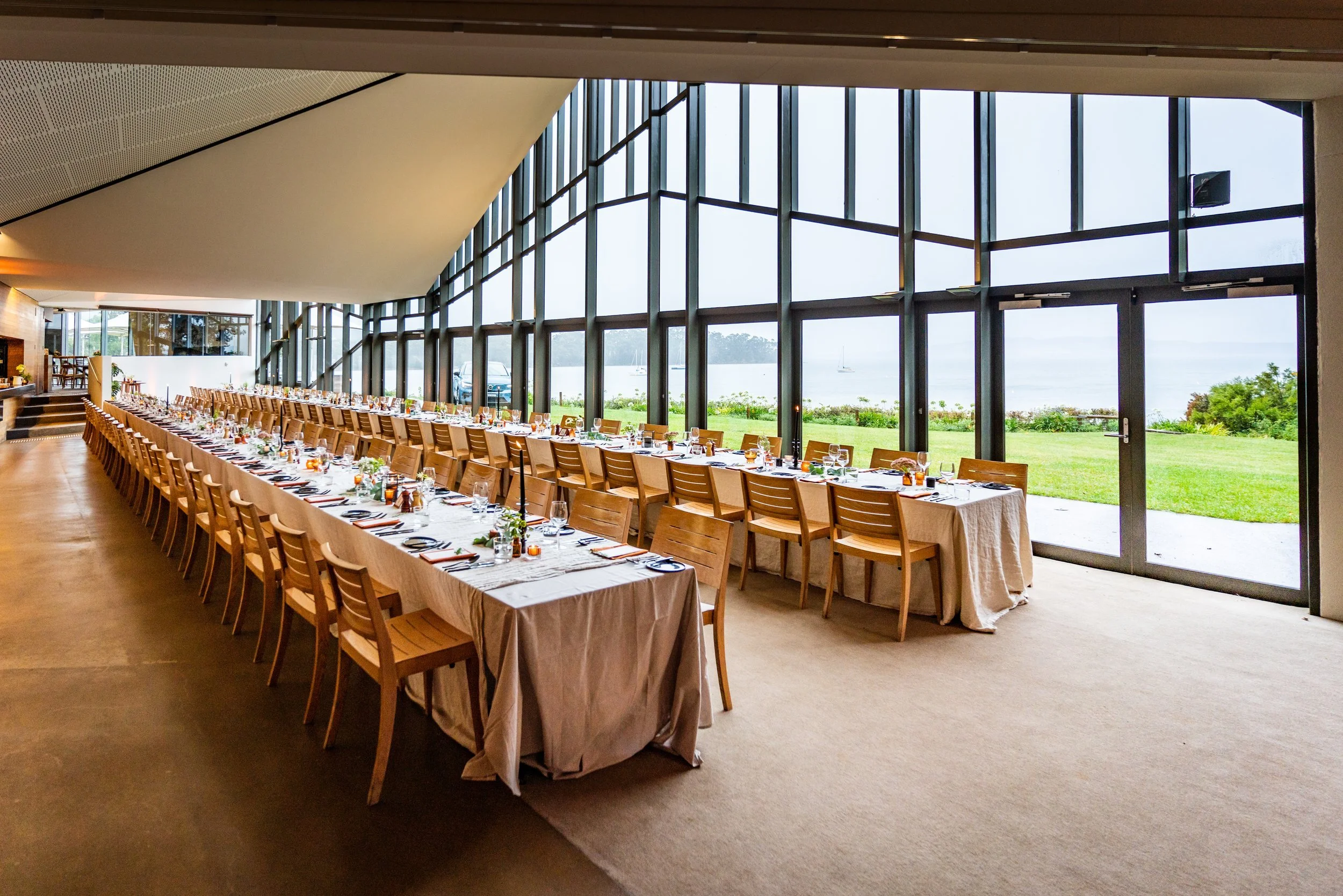 Interior of a banquet hall with a long table set up for a meal, with glassware and plates, overlooking a large window with a view of a lake and sailboats outside.