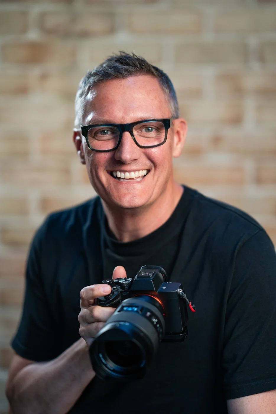 A man with glasses holding a professional camera and smiling, against a blurred brick wall background.