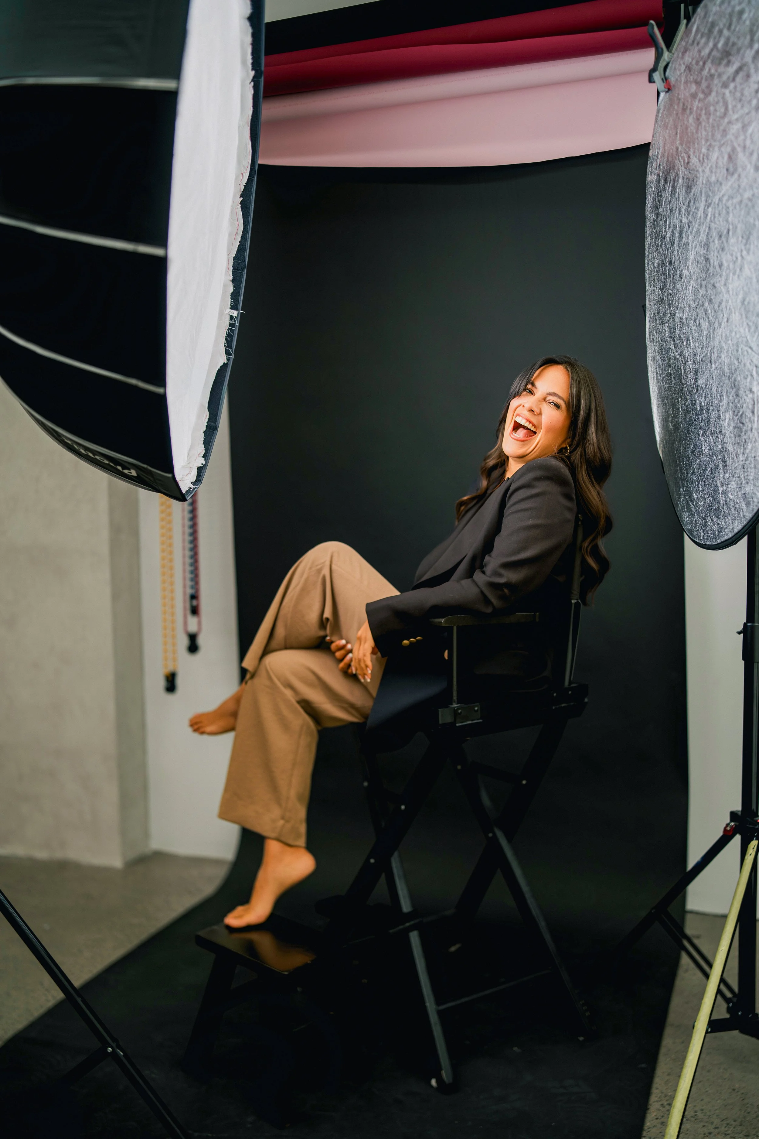 A woman sitting on a director's chair, laughing with her mouth open, in a photoshoot studio with black backdrop and professional lighting.