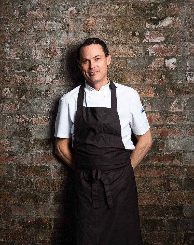 Professional chef standing against a brick wall, wearing a white chef's coat and a black apron.