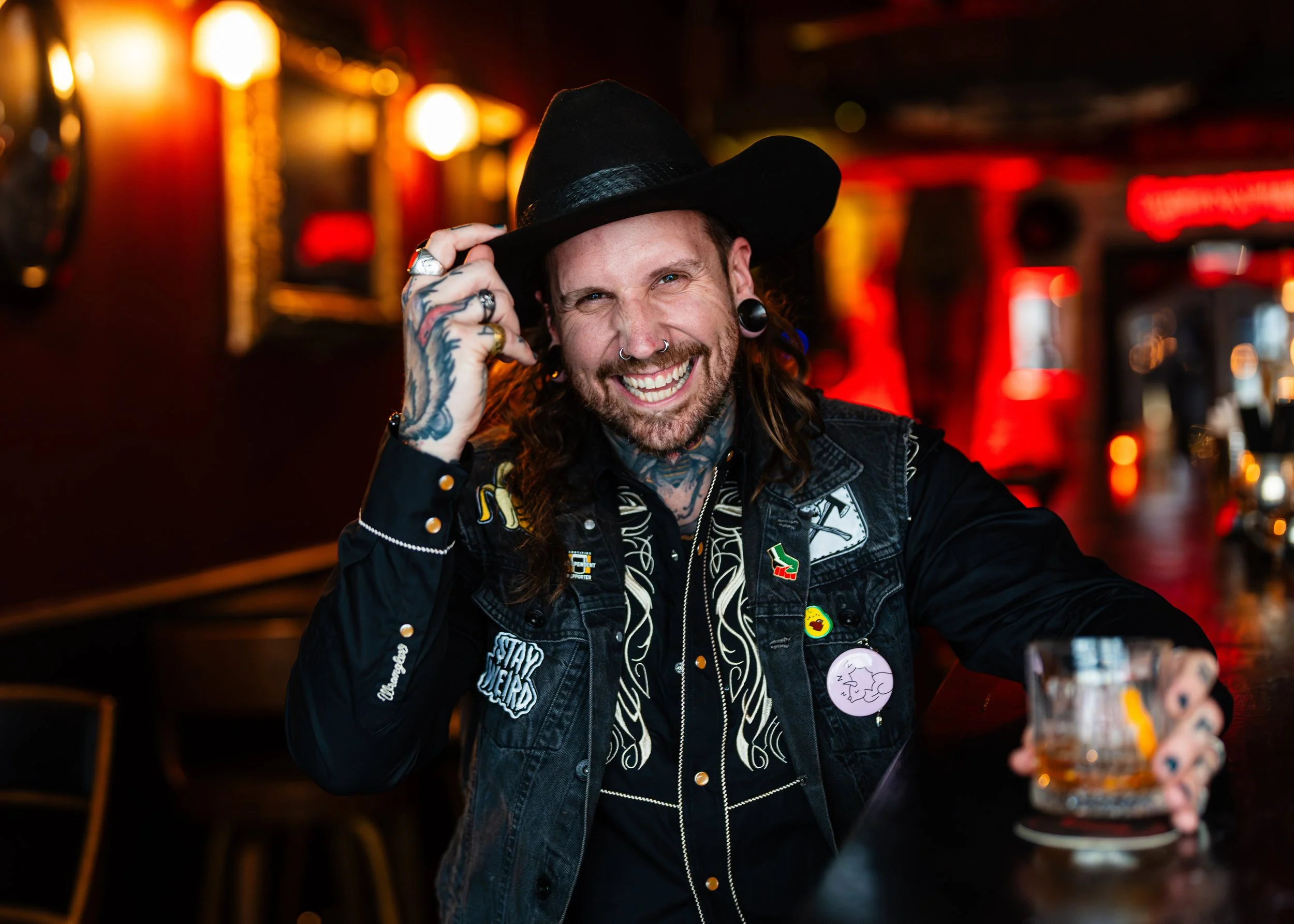 A smiling man with tattoos, piercings, and long hair, wearing a black cowboy hat and a black vest with patches, sitting at a bar in a dimly lit venue with red and yellow lights.