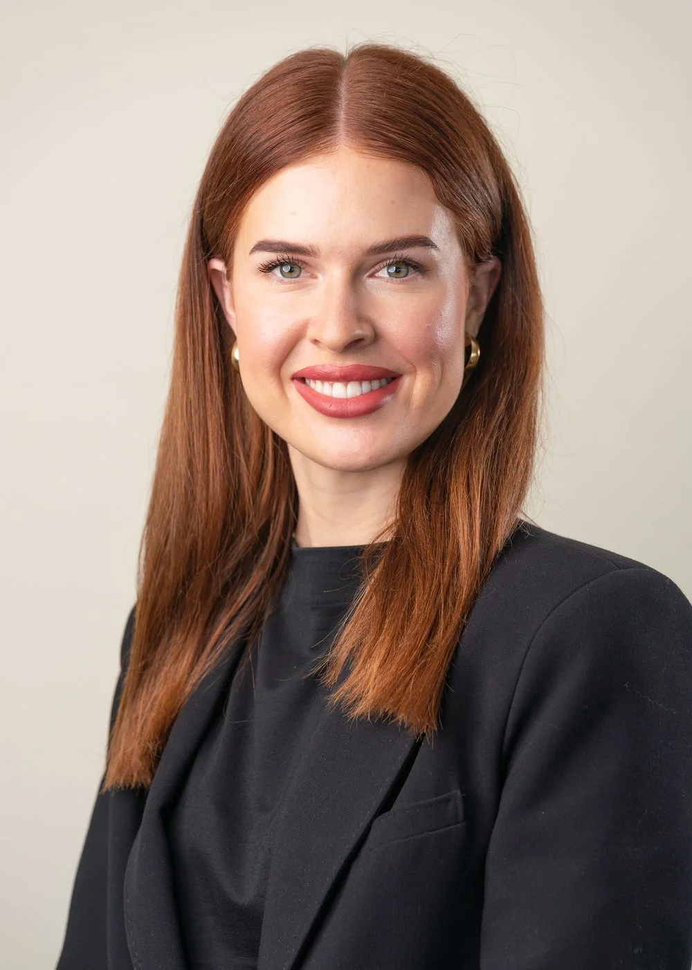 A portrait of a woman with long red hair, wearing a black blazer and a black top, smiling and looking at the camera against a plain light background.