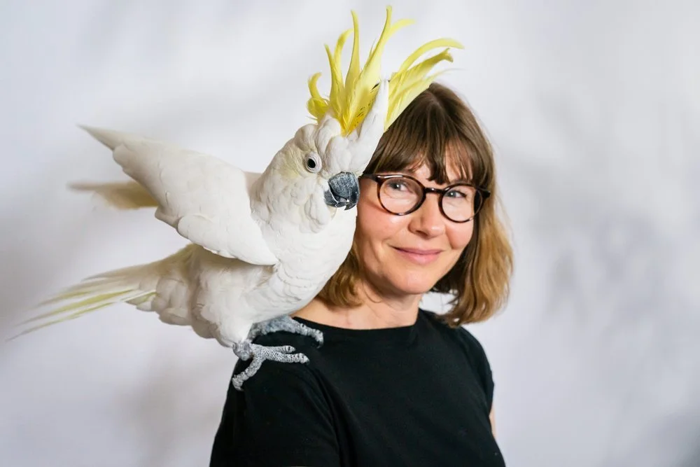 A woman with glasses and shoulder-length brown hair smiling while looking at the camera, with a white cockatoo on her shoulder. The cockatoo has a yellow crest and appears to be blowing a kiss.