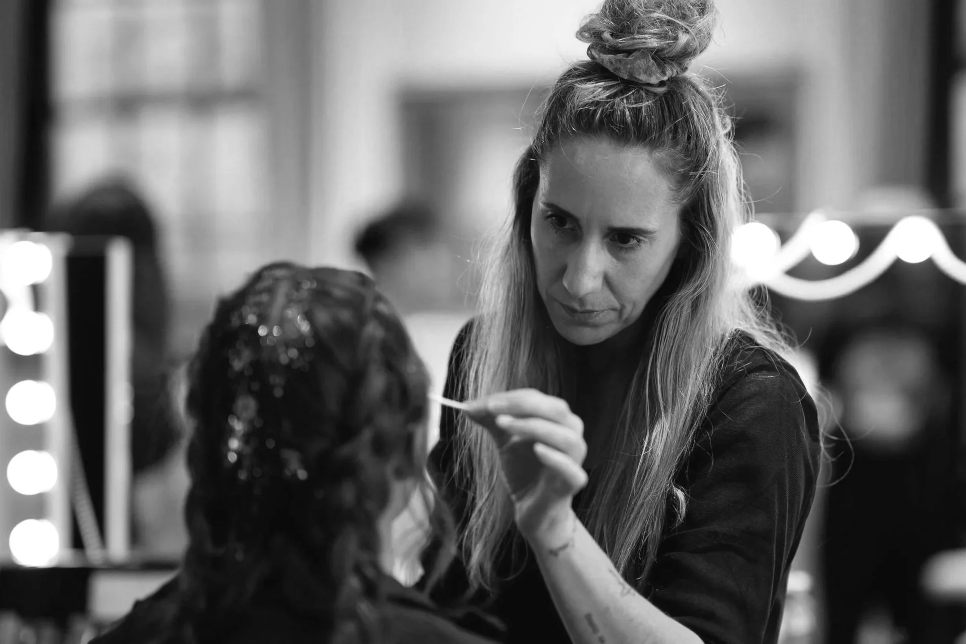 A woman with long hair styled in a top bun is applying makeup or face paint to a person's face. The scene appears to be in a backstage or dressing room with lights and mirrors in the background.