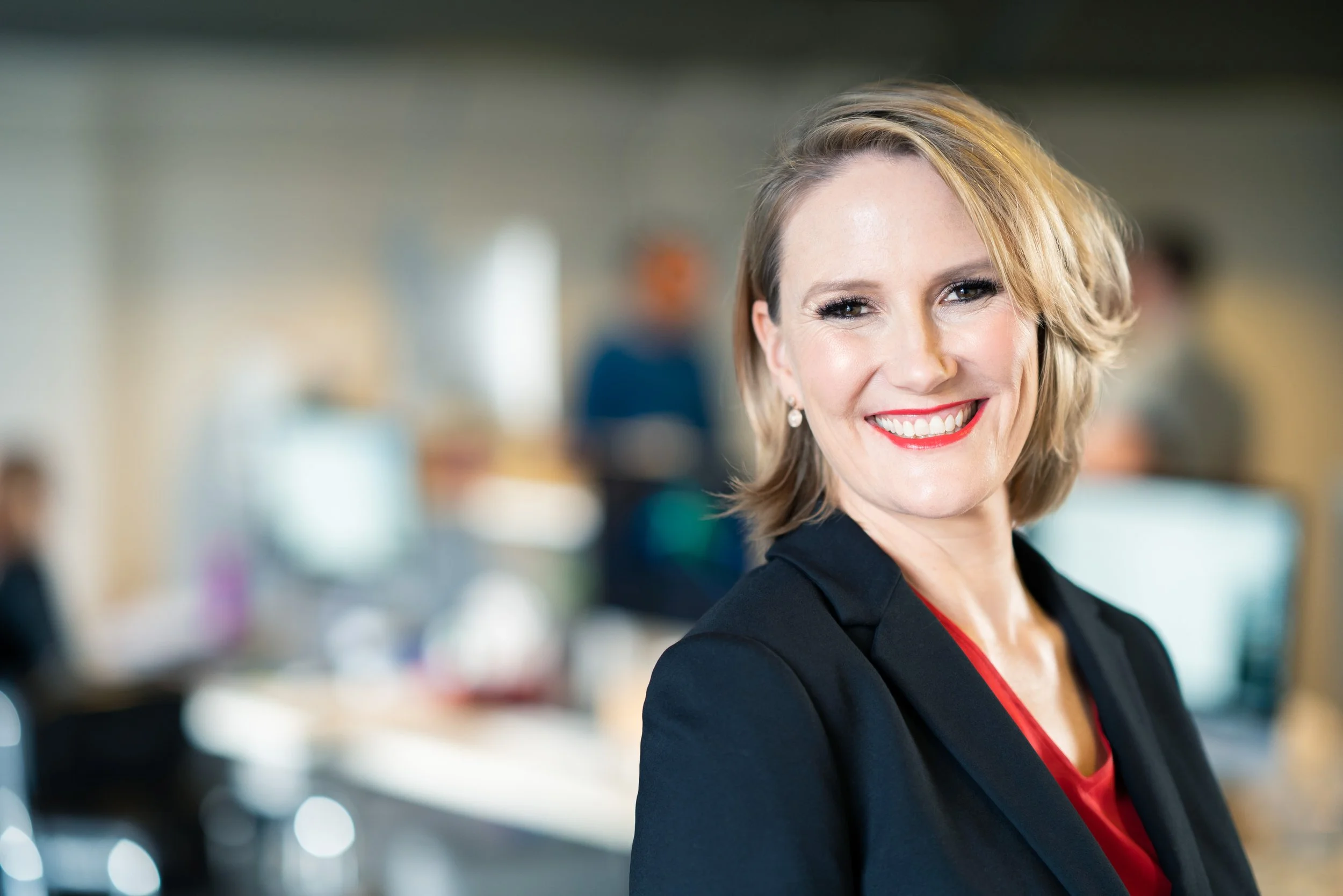 Smiling woman with blonde hair and red lipstick in professional attire, standing in an indoor environment.