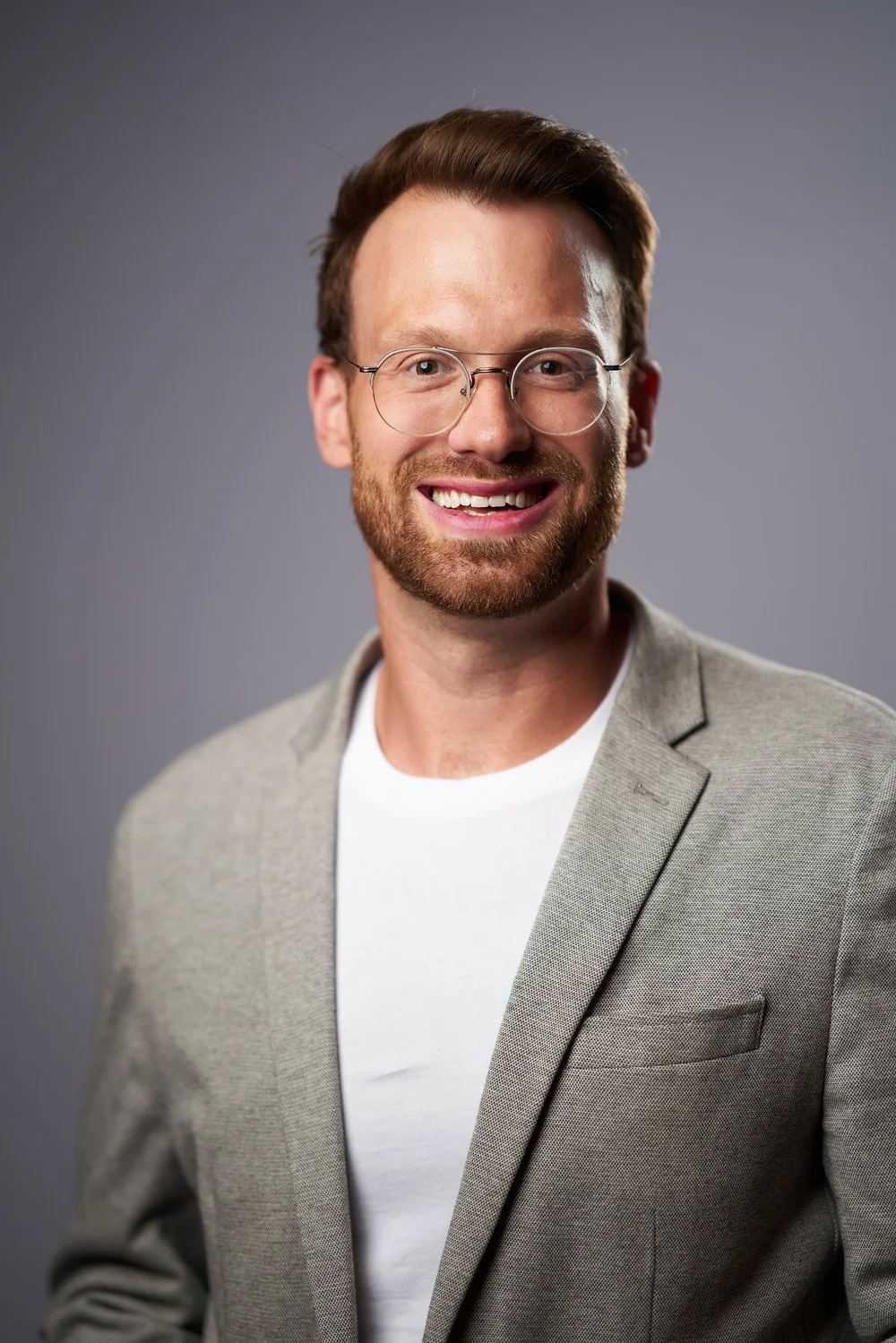 Portrait of a smiling man with glasses, ginger hair, and a beard, wearing a light gray blazer over a white t-shirt, against a neutral gray background.