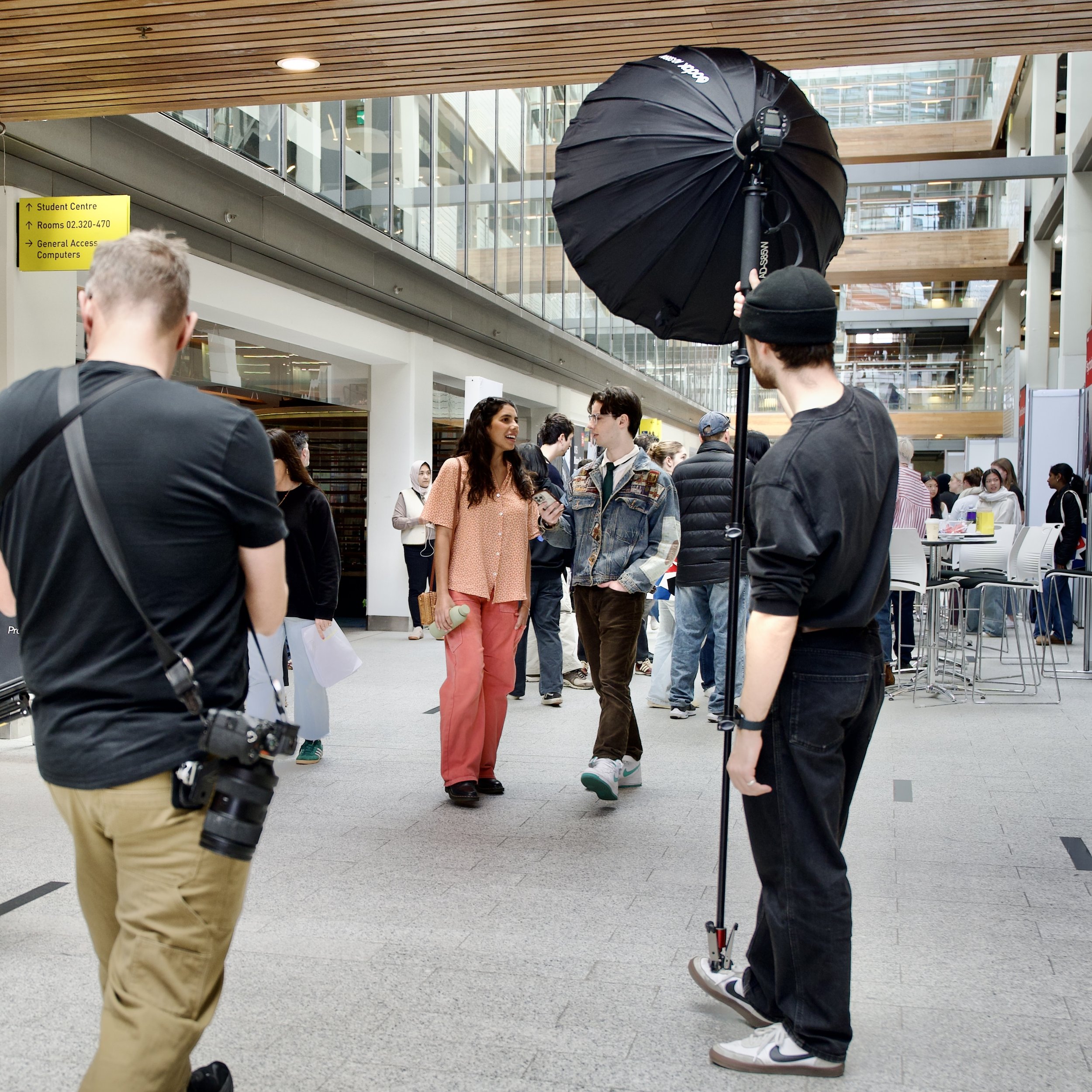 Group of people inside a modern building with large windows, some engaging in conversation, and a studio lighting setup with a black umbrella for a photoshoot.