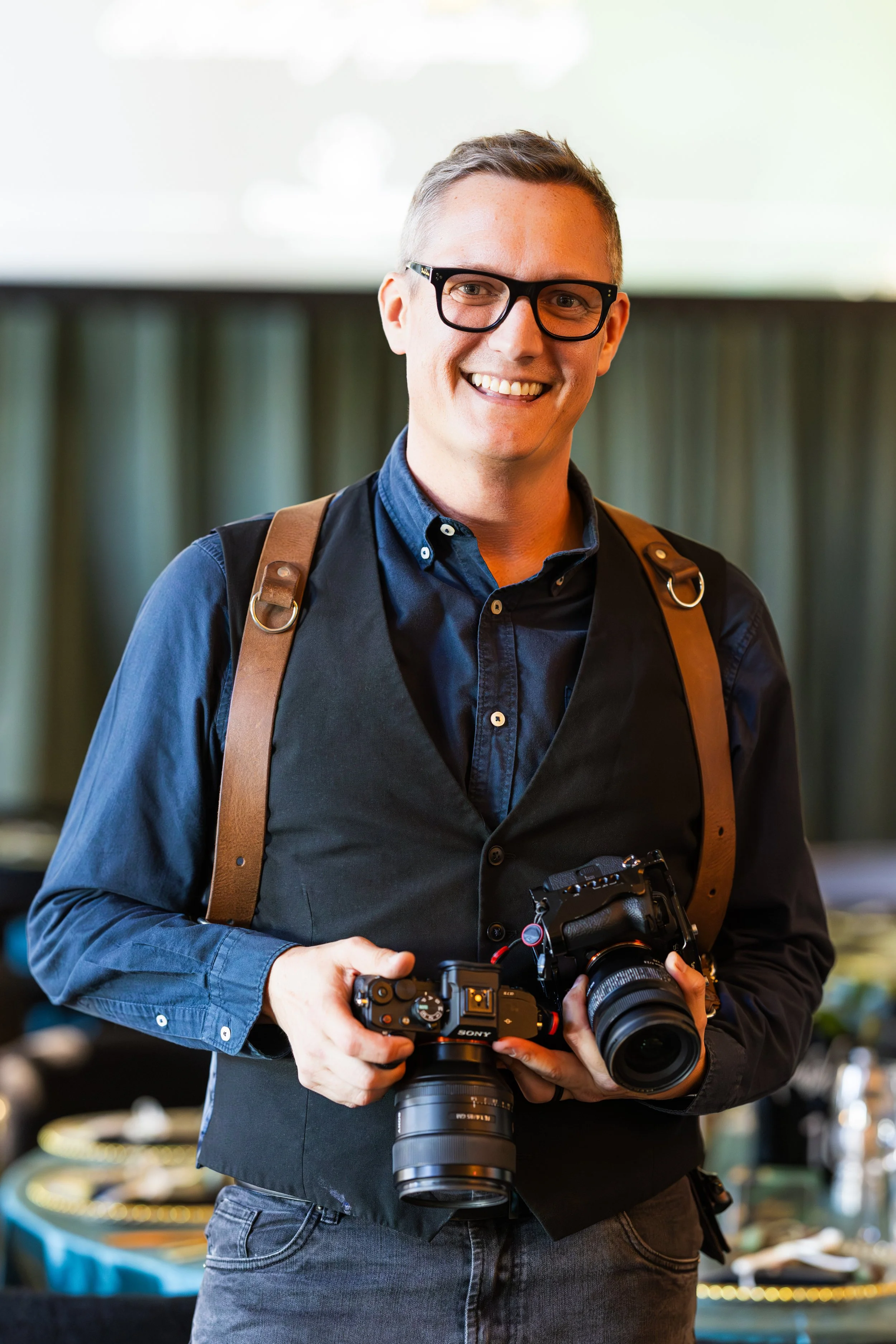 A smiling man wearing glasses, a dark shirt, and a vest, holding two professional cameras in an indoor setting with a blurred background.