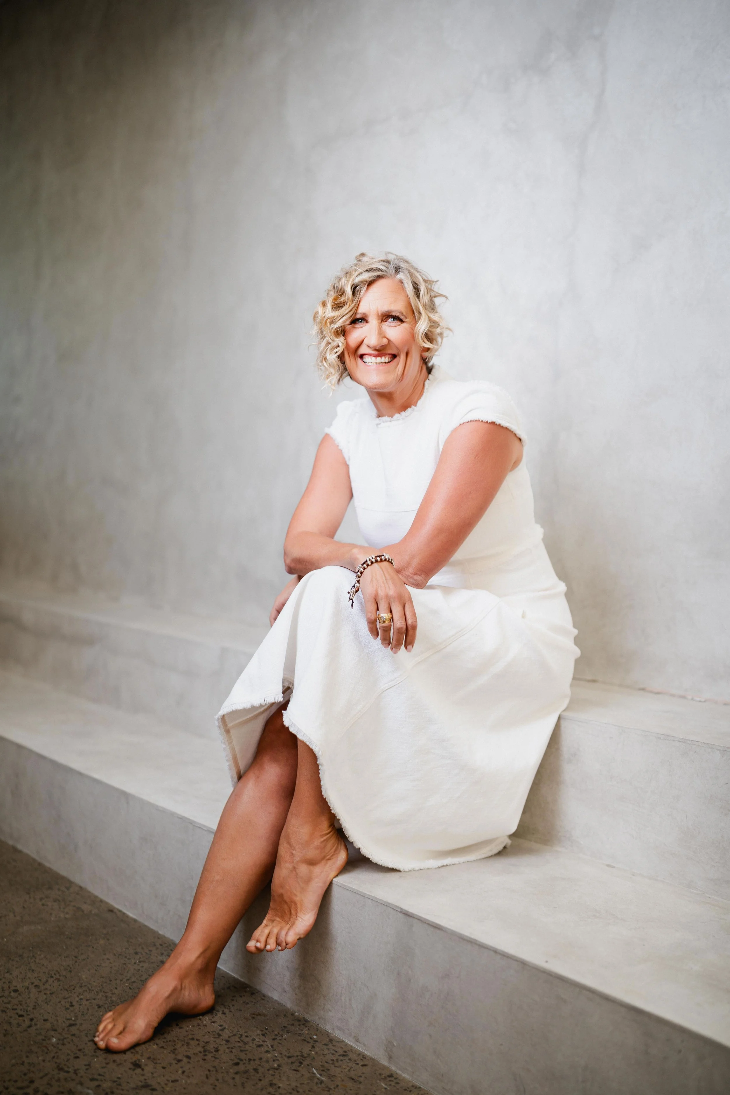 A woman with curly blonde hair sitting on a white concrete step against a light gray wall, smiling and wearing a white dress, with her legs crossed and barefoot.