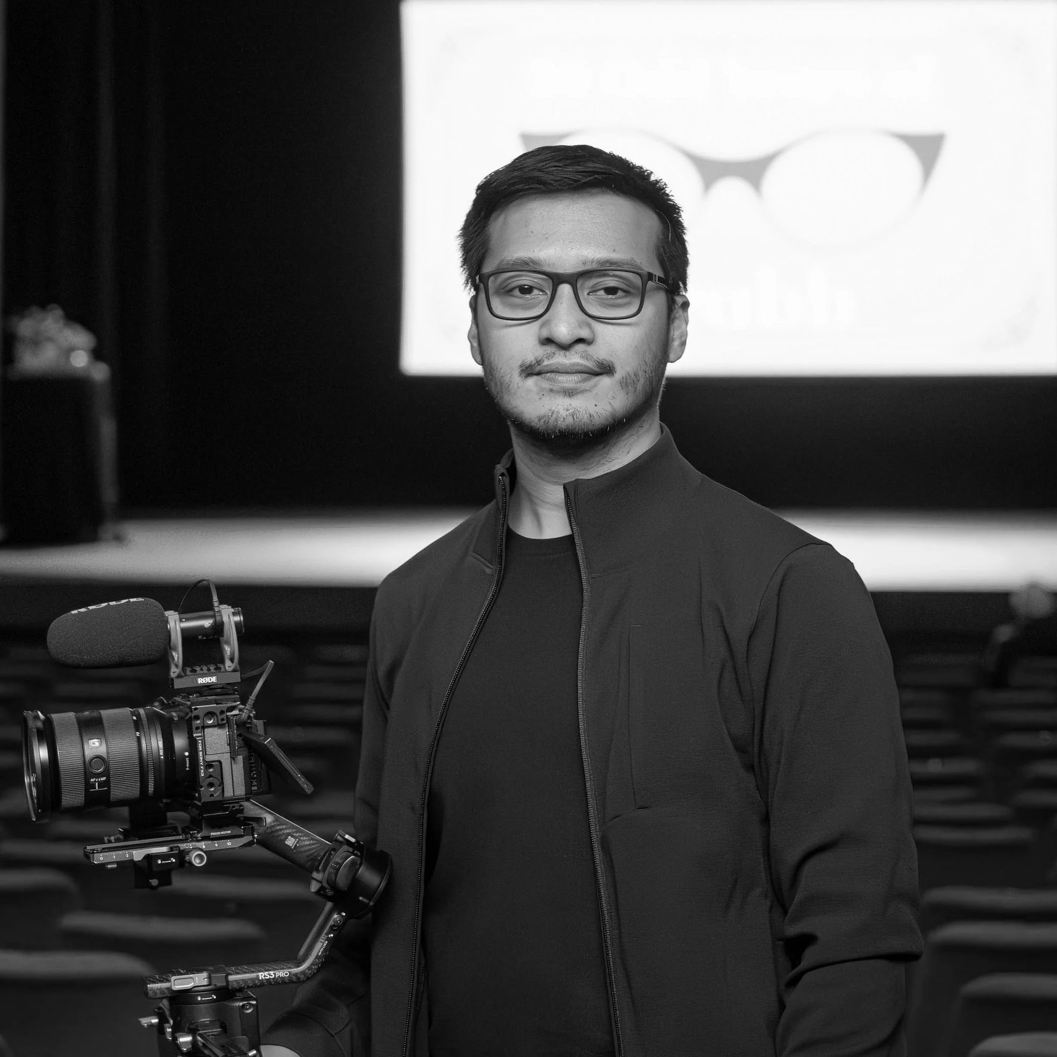 A man with glasses and facial hair holds a professional camera mounted on a stabilizer, standing in front of a bright screen with a logo in the background.