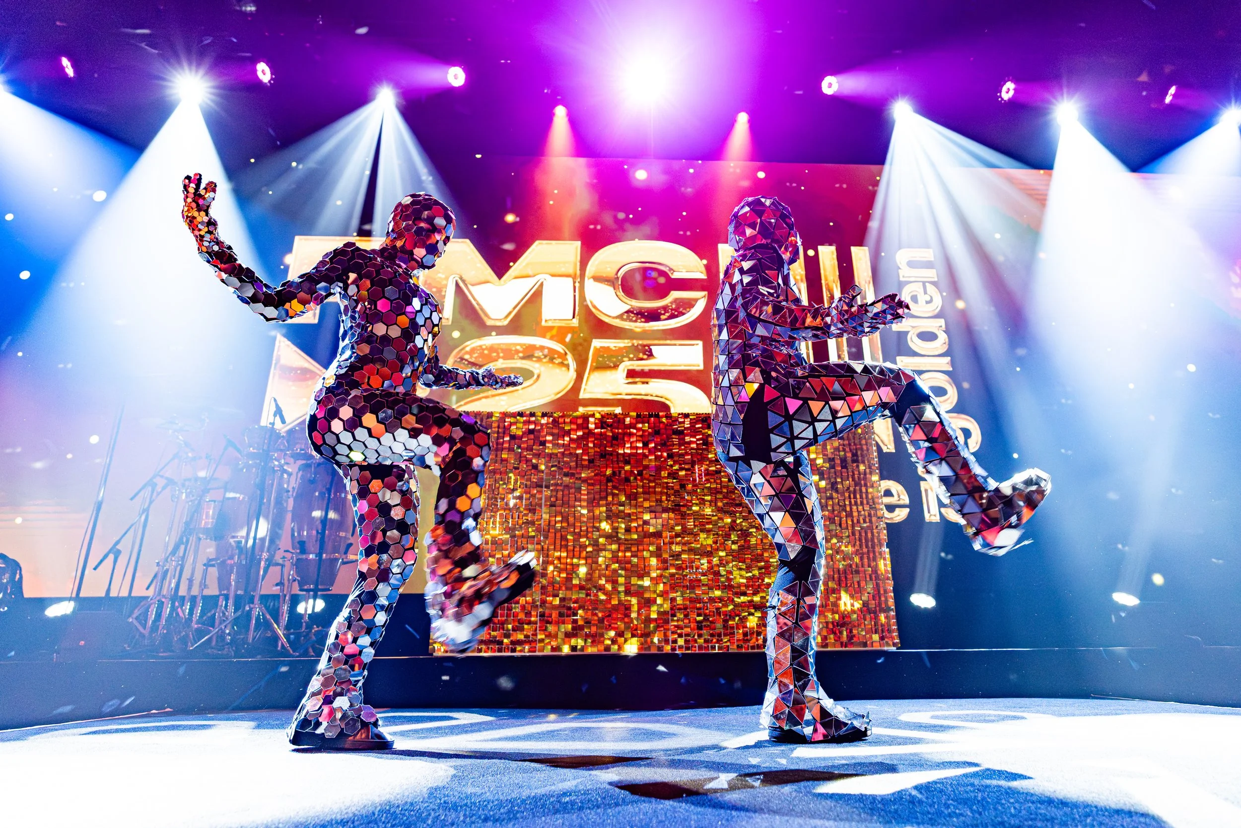 Two dancers made of reflective, mosaic-like material perform on stage with bright colorful lighting, a large sign with the letters 'MCAT' in the background.