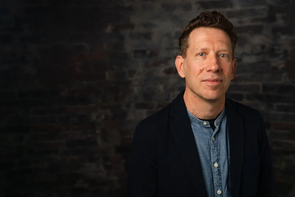 A man with short curly brown hair and light skin, wearing a blue shirt and dark jacket, stands against a dark brick wall.