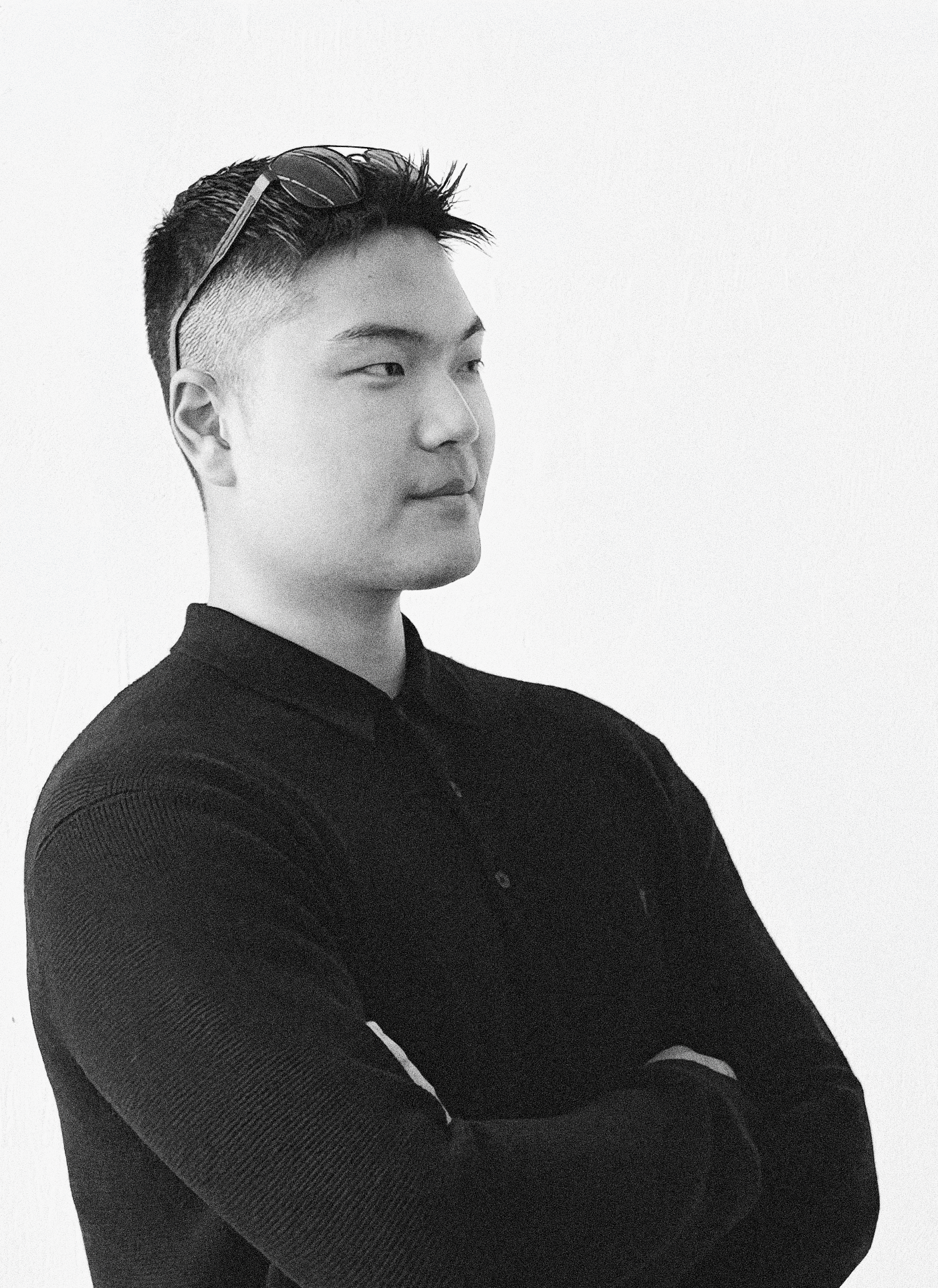 A black and white photo of a young man with short, styled hair, glasses resting on his head, wearing a black long-sleeve shirt with a textured pattern, standing with arms crossed against a plain white background.
