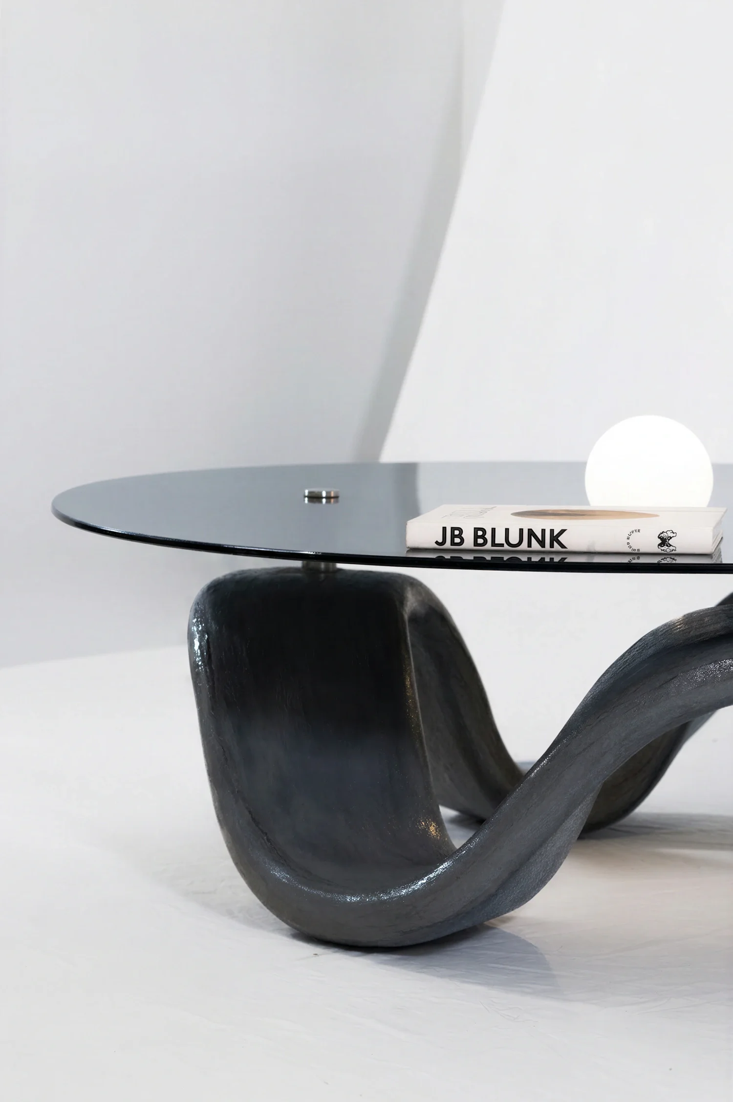 A modern black chair with a rounded, sculptural wooden base, placed next to a black round side table with a clear glass top, on which a book titled 'JB BLUNK' rests.