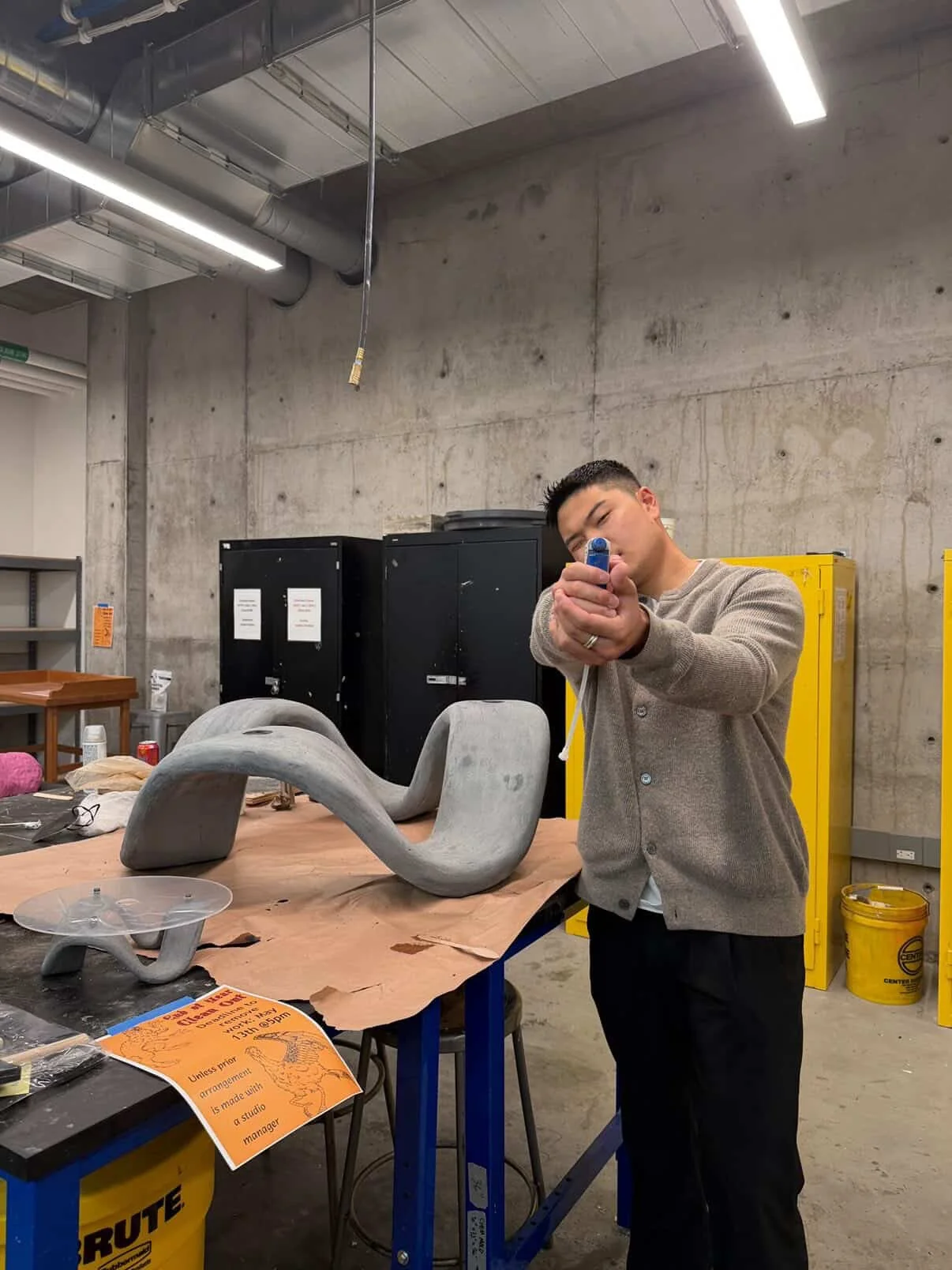 A man in a gray sweater aiming a small blue object, possibly a miniature sculpture, in a workshop with concrete walls, black cabinets, a yellow locker, and a table with art supplies. There is a large, abstract gray sculpture on the table.