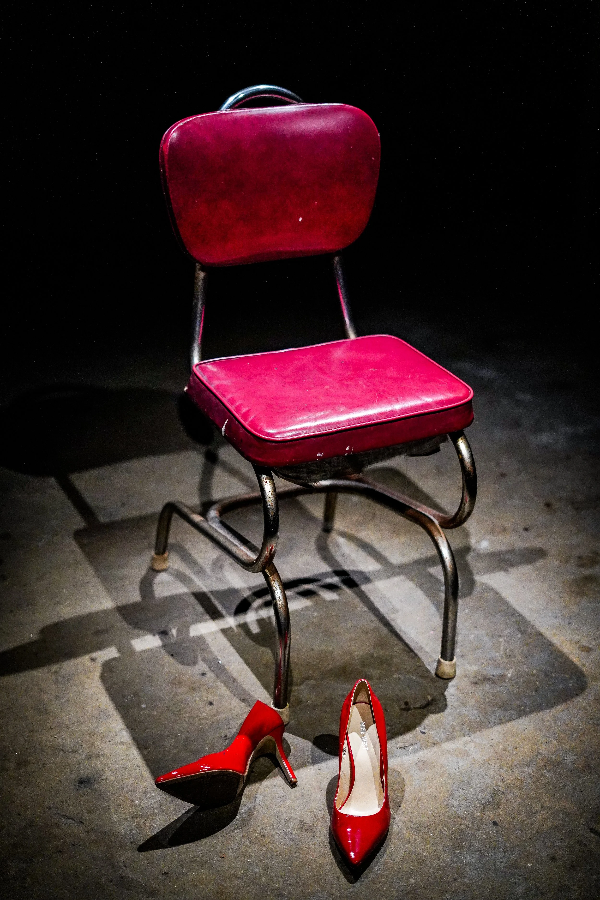A vintage red chair with a shiny seat and backrest, with a pair of red high-heeled shoes placed in front of it on a concrete floor, casting shadows.
