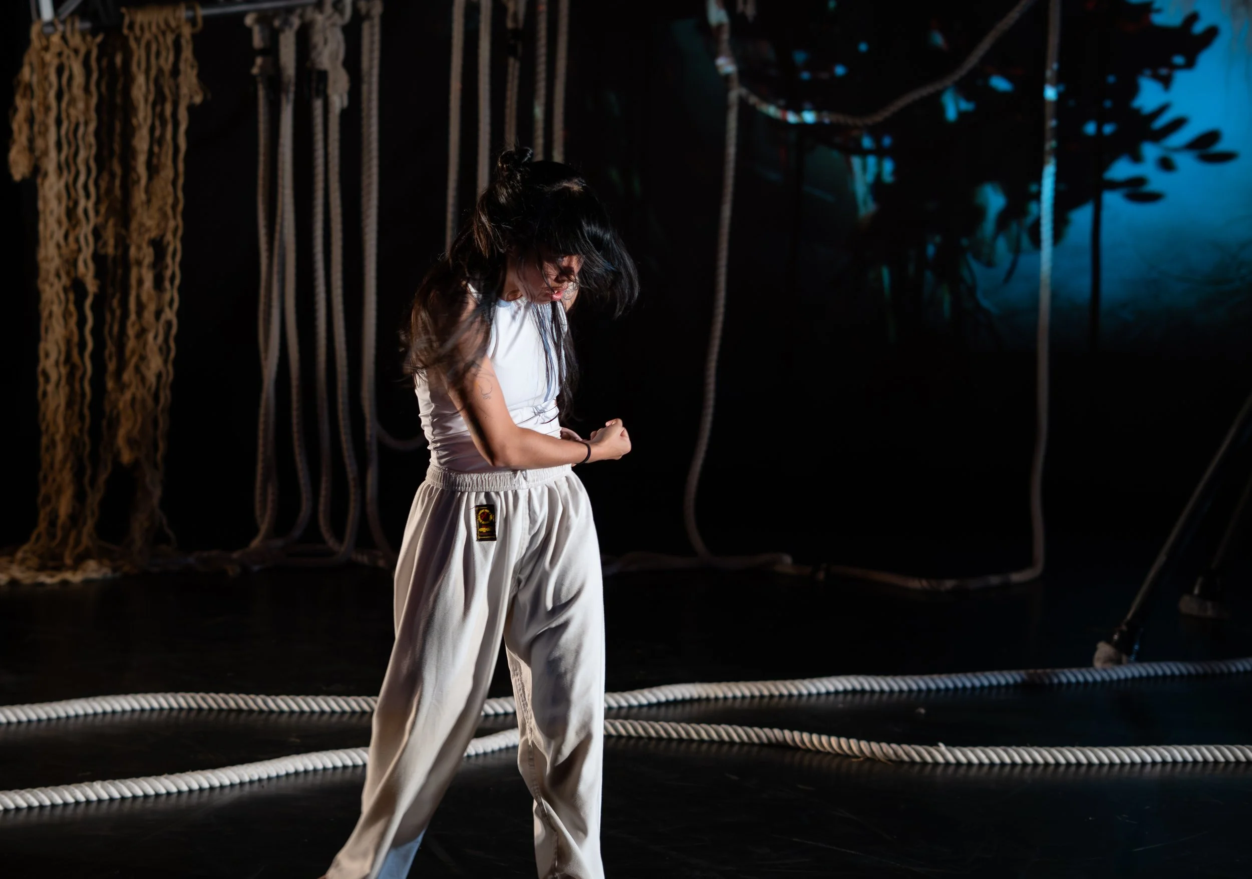 A young woman with long dark hair wearing a white sports bra and loose white pants standing in a dimly lit room with ropes hanging from the ceiling.