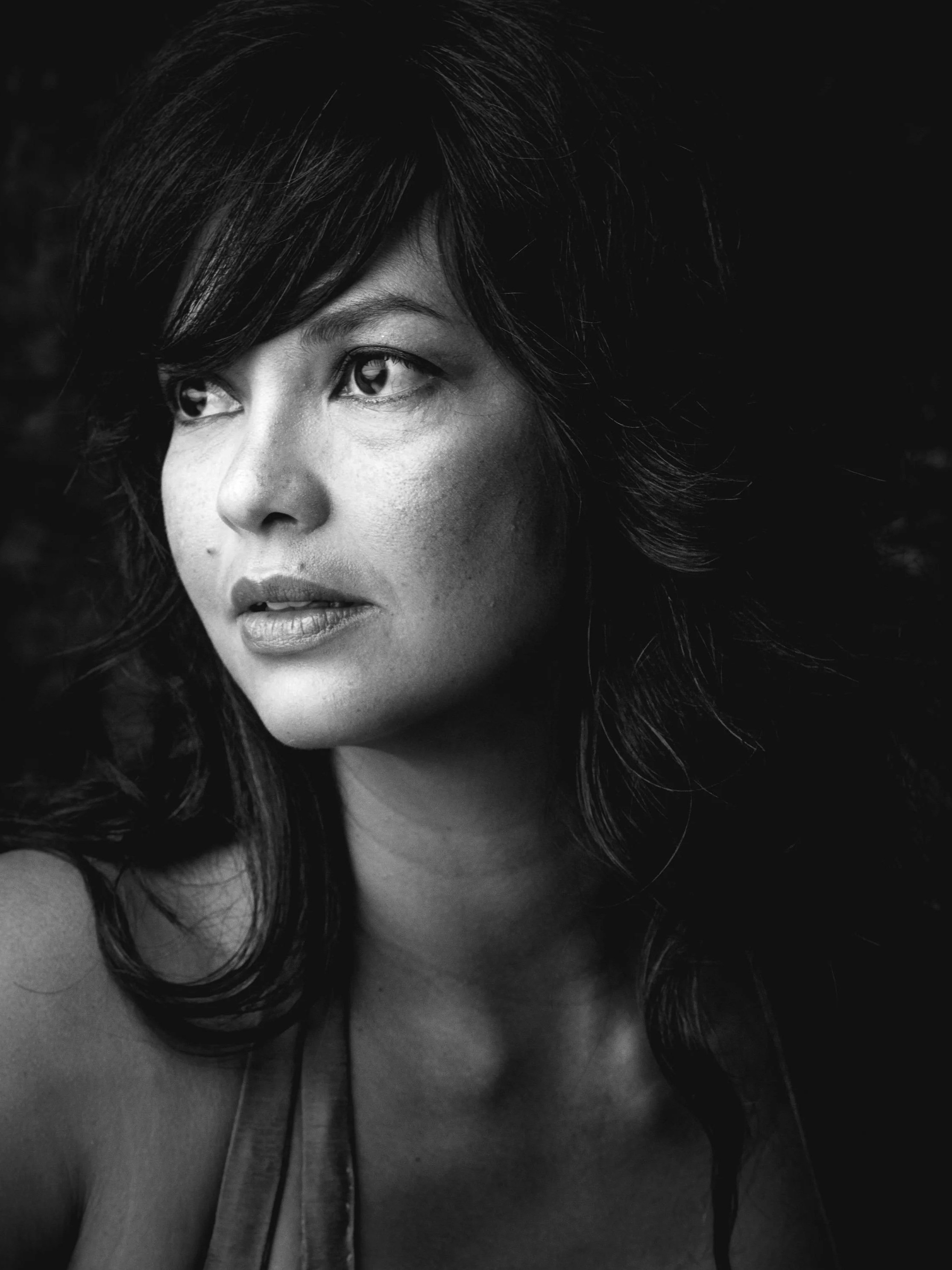 A black and white close-up photograph of a woman with dark, wavy hair and smooth skin. She is looking slightly upward and to the side with subtle makeup, including lipstick and eyeliner. The background is dark and out of focus.