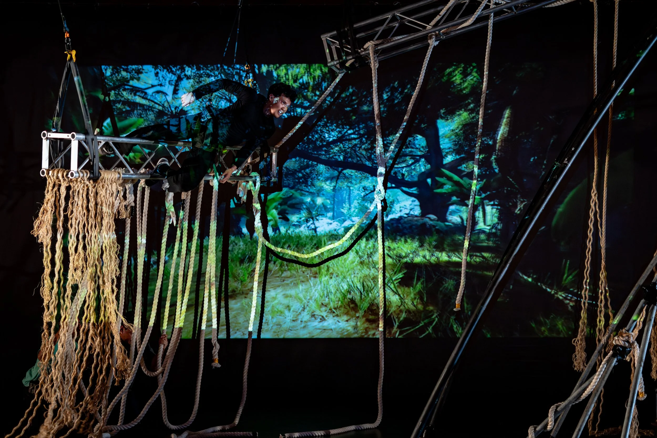 A performer crawling on a platform suspended by ropes against a backdrop of a forest scene.