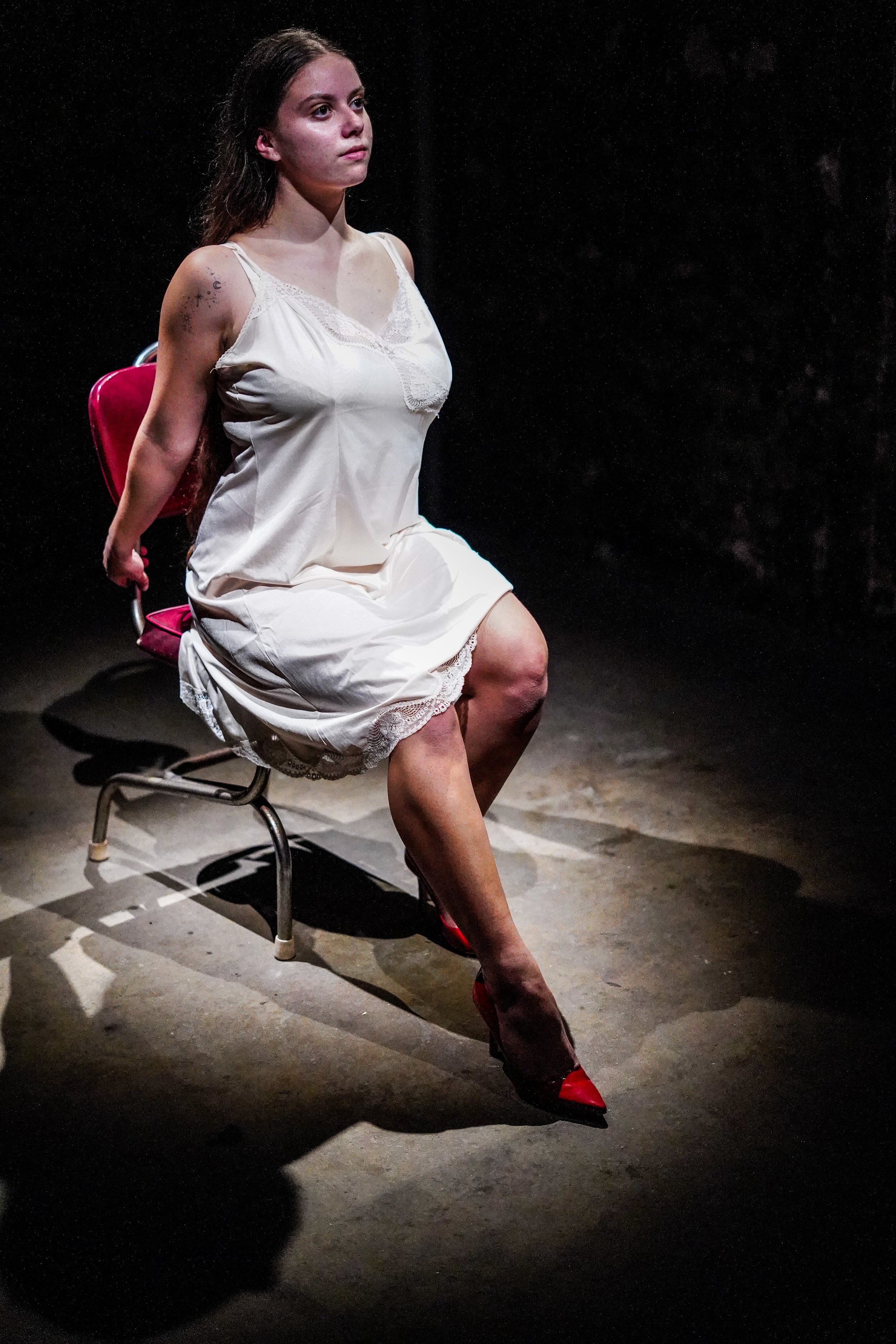 A woman with long hair, wearing a white nightgown and red heels, is sitting on a small chair in a dark, empty space with a black background. She appears to be in a dramatic or contemplative pose.