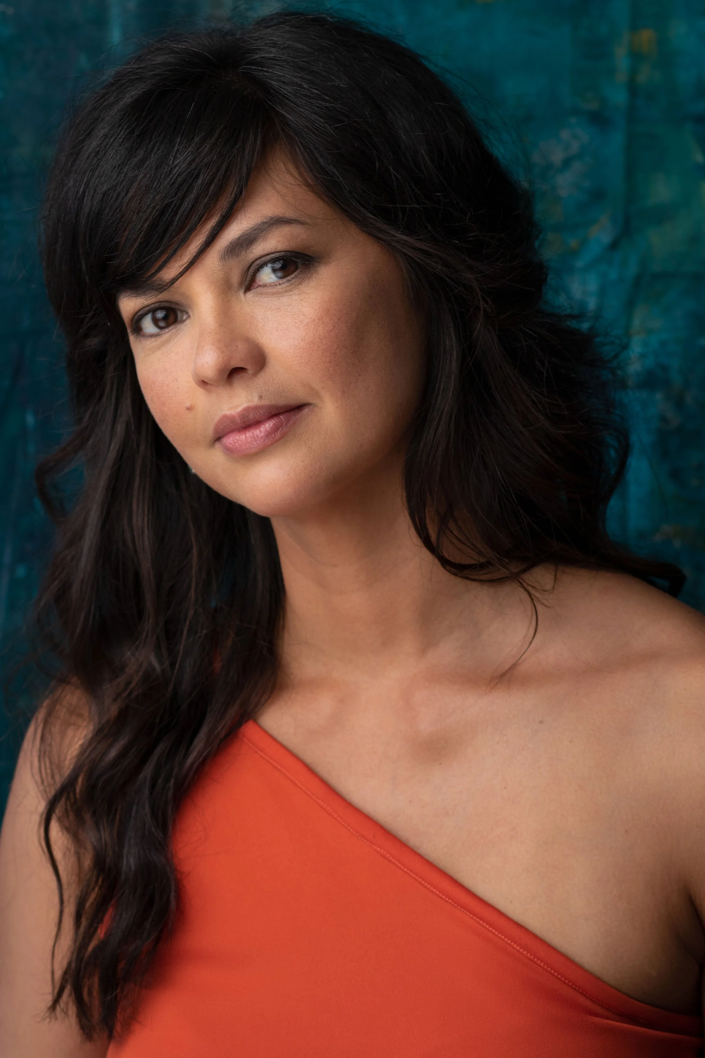 A woman with dark, wavy hair, wearing an orange dress, looking at the camera with a slight smile. The background is textured blue.